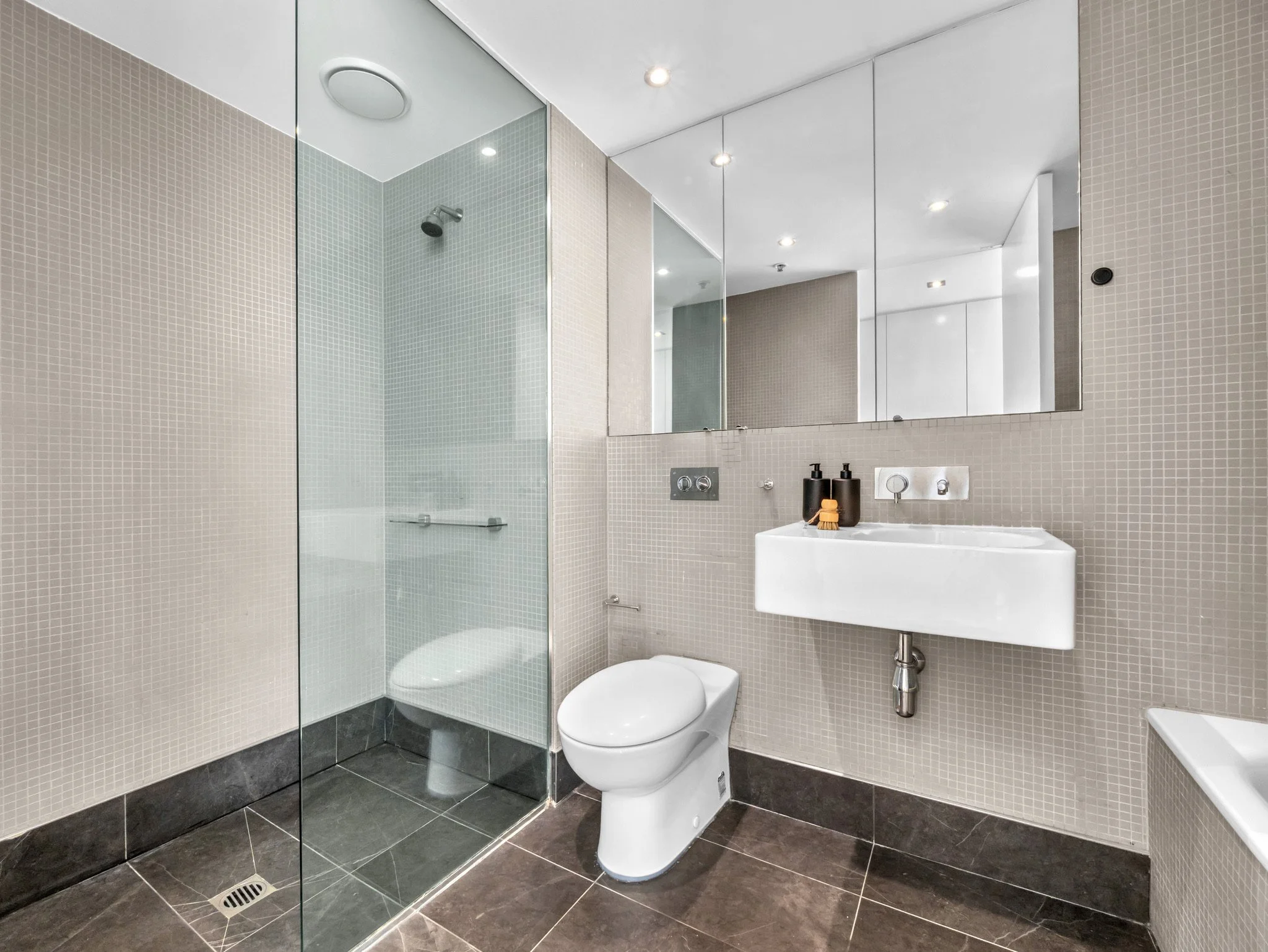 A modern bathroom with beige small tile walls, dark tiled floor, a glass-enclosed shower, a white toilet, a white sink with black pump soap dispenser and a black pump bottle, and a large mirror on the wall.