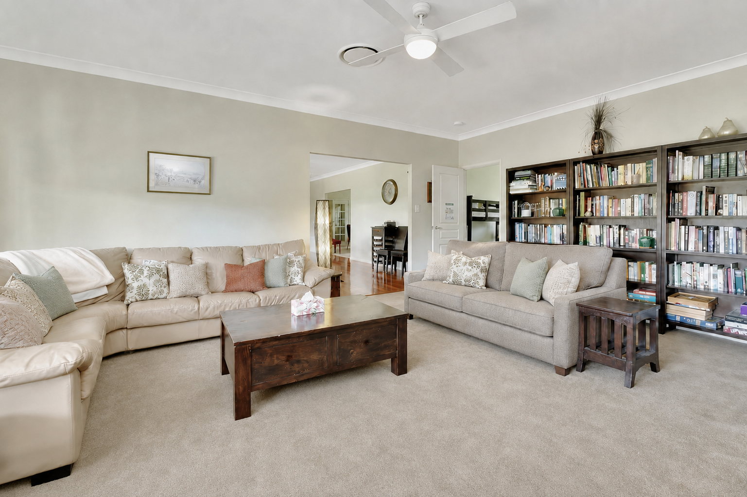 Living room with beige sectional sofa and gray sofa, bookshelf filled with books, wooden coffee table, and beige carpet.