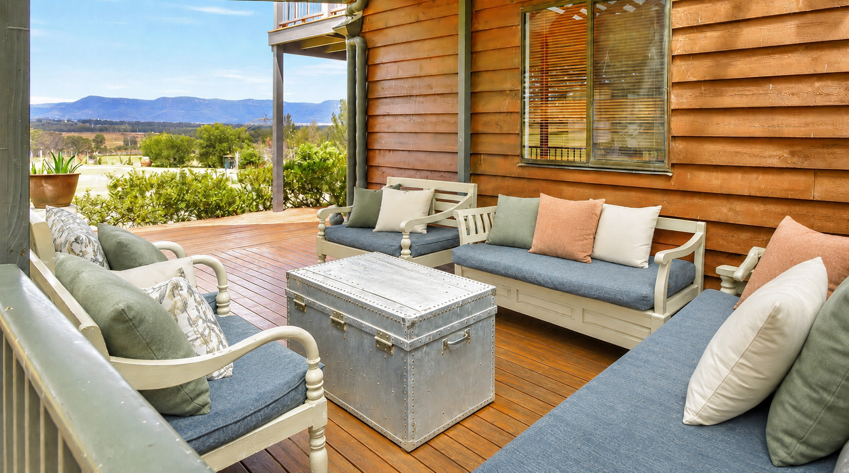 Outdoor patio with wooden deck, beige and blue cushioned benches and chairs with pillows, metal storage trunk, green bushes, distant mountains, and a cloudy sky.
