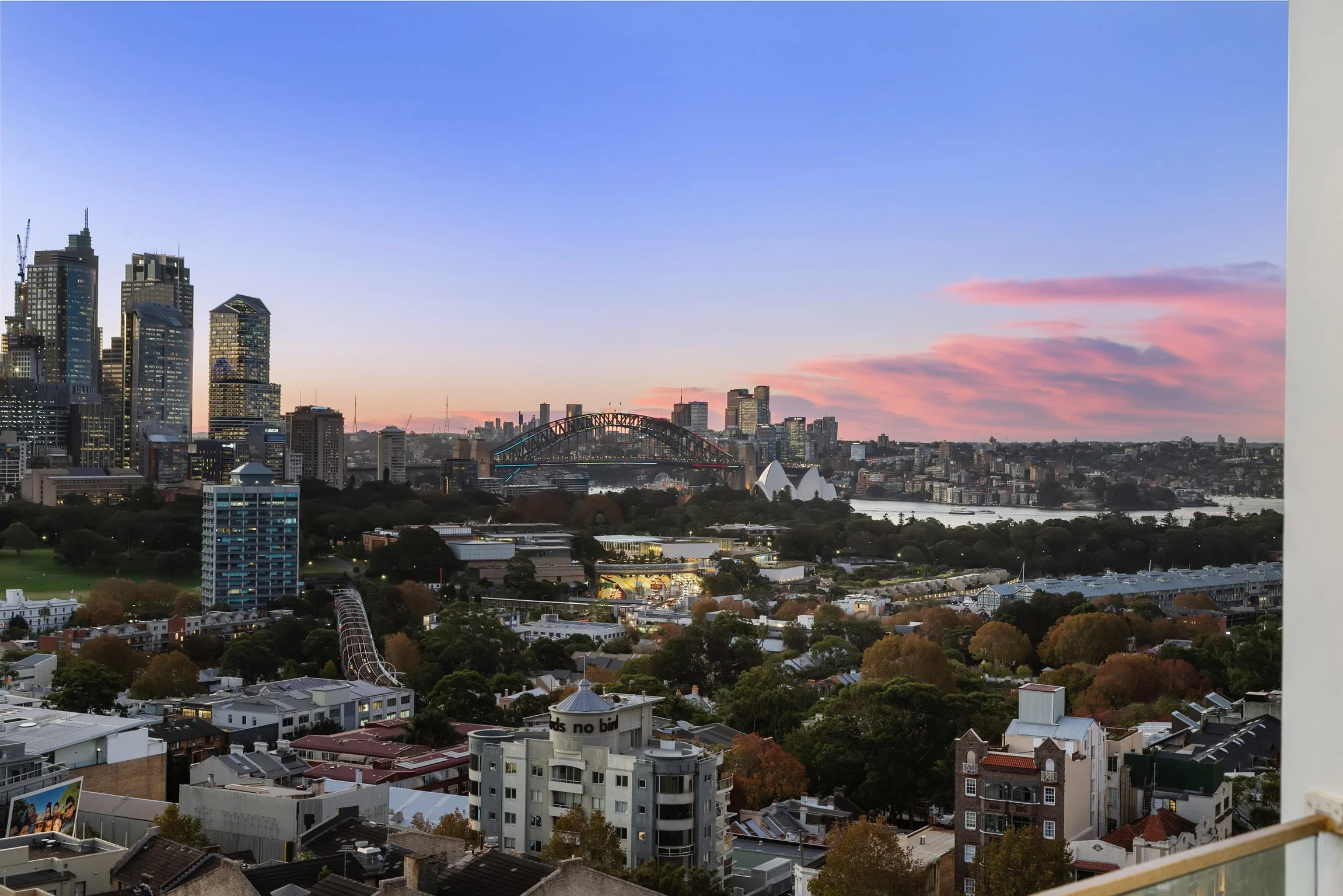 City skyline at sunset with tall skyscrapers, an arched bridge, and a river with pink clouds in the sky.