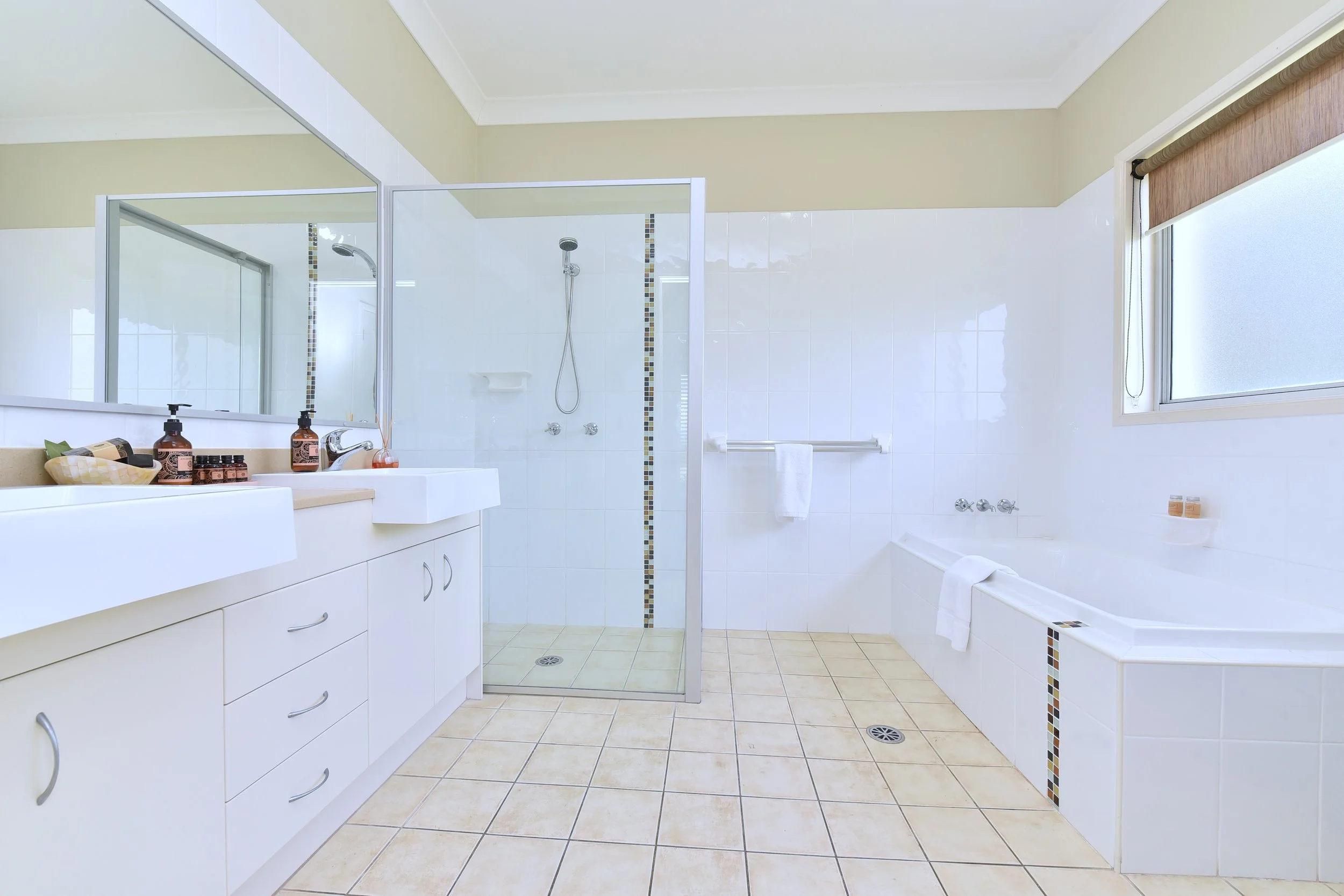 A clean, bright bathroom with a white vanity, a large mirror, a glass shower enclosure, a bathtub, and a frosted window with a wooden roller blind.