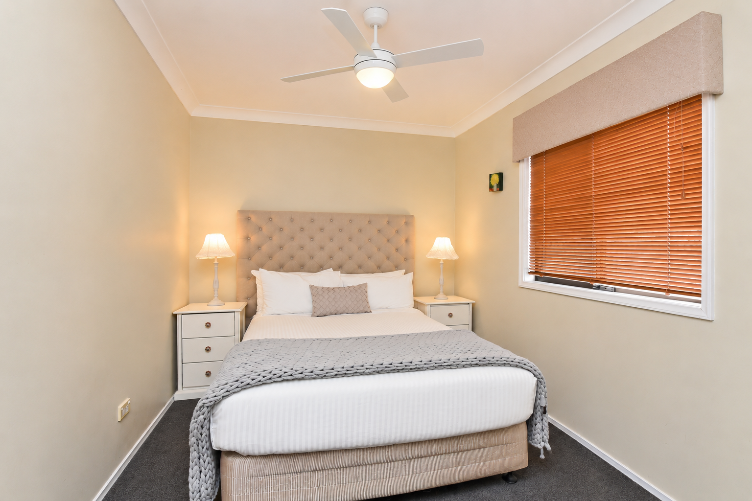 A bedroom with a beige upholstered bed, beige walls, white nightstands, and table lamps. There are window blinds and a ceiling fan.