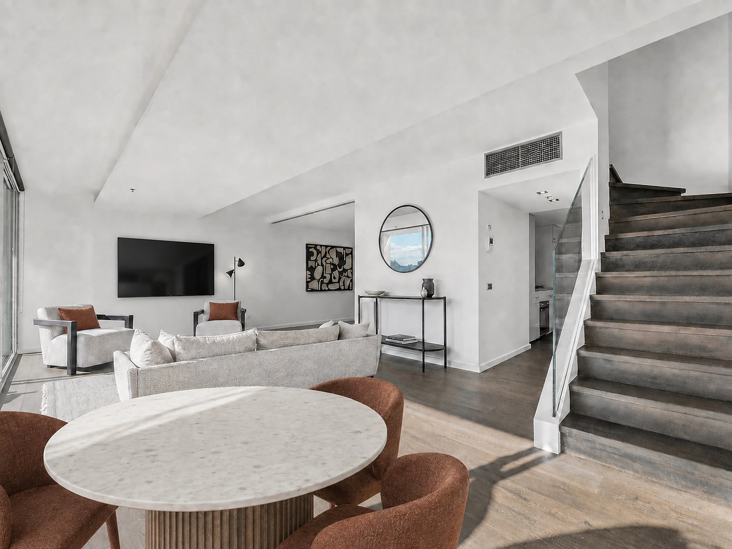 Modern living room with white walls, a large flat-screen TV, a white sofa, orange and white chairs, round dining table, staircase with dark wood steps, and decorative wall art.