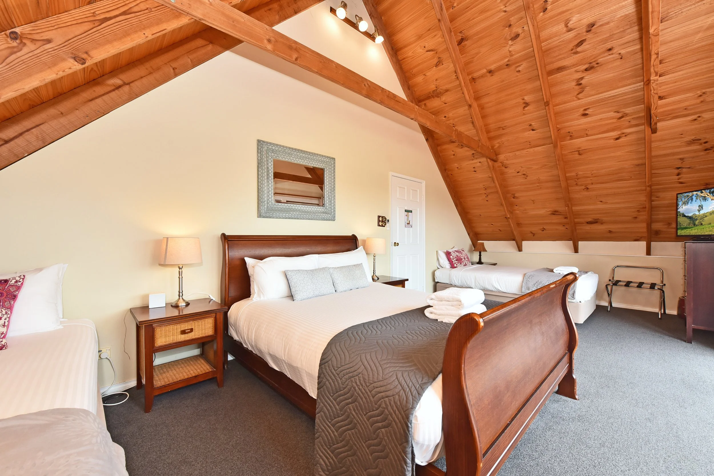 Bedroom with two beds, wooden ceiling with beams, lamps on nightstands, mirror above one bed, TV in corner, and towels on the bed.