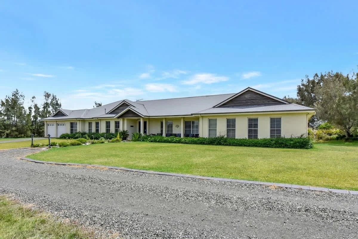 A large, single-story house with a light yellow exterior, gray roof, and multiple windows is shown in a spacious yard with green grass and landscaping. There is a gravel driveway in front of the house and trees in the background under a blue sky.