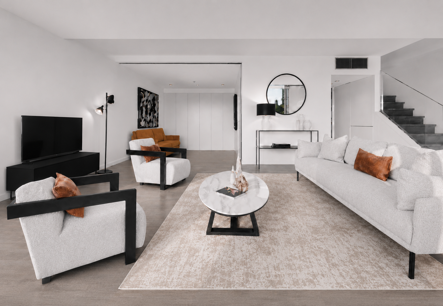 Modern living room with white walls, black and white furniture, a beige rug, and a staircase. Features include a large white sofa with brown cushions, a white armchair, a black TV on a black stand, and a marble coffee table with decorative vases.