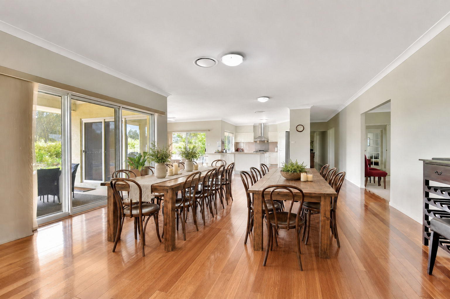 Open-concept dining area with wooden tables and chairs, decorated with candles and plants, adjacent to a kitchen with white cabinets and stainless steel appliances. Large windows and a sliding glass door lead to an outdoor patio.