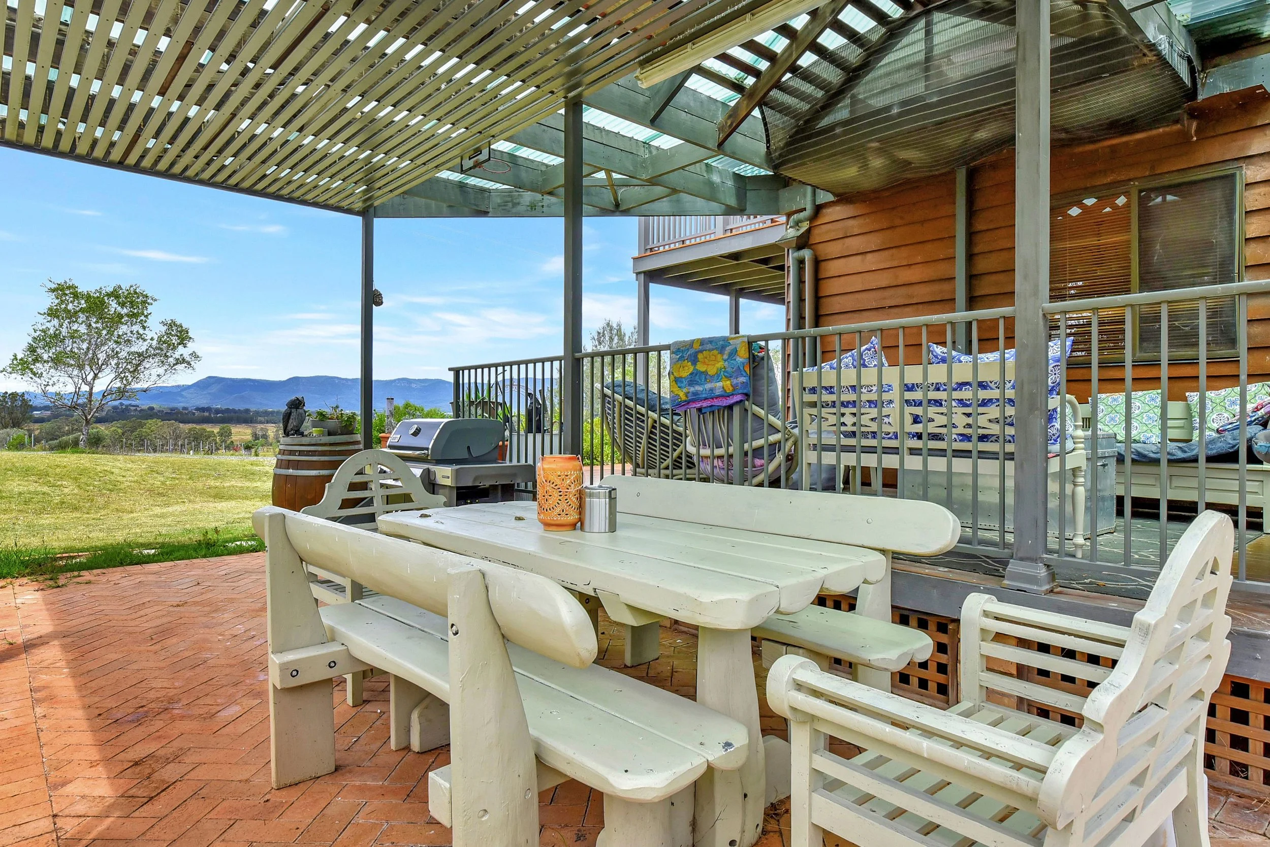 A covered outdoor patio with a white picnic table and benches, a barbecue grill, and various chairs. The area has a view of distant mountains and a grassy field, with some trees and a partly cloudy sky in the background.