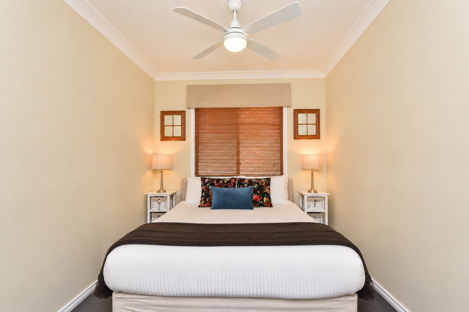A bedroom with a white bed, dark brown blanket, and decorative pillows, flanked by white nightstands with lamps. Windows with blinds and wooden frames are behind the bed, and a ceiling fan is above.