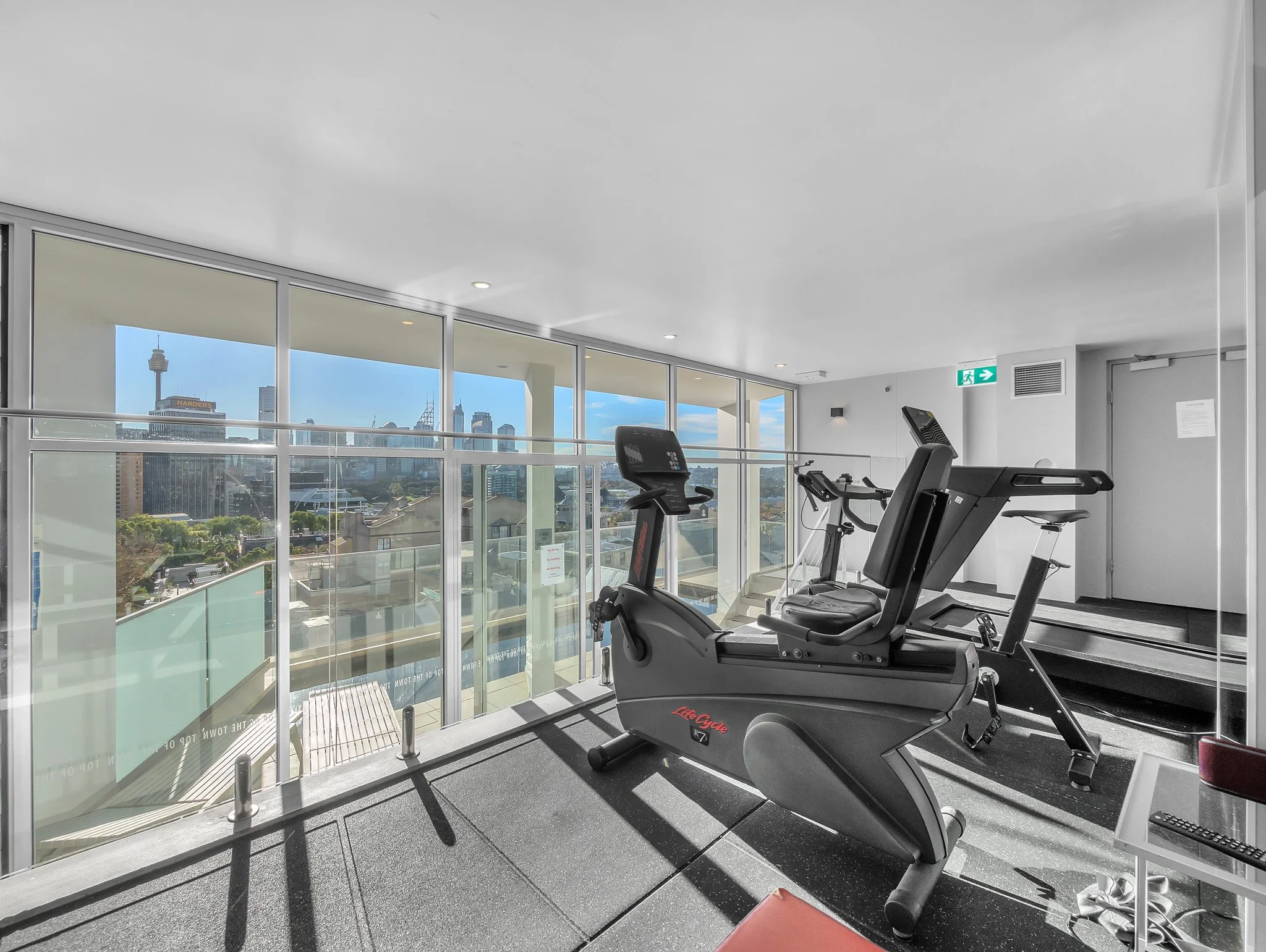 Fitness center with elliptical machines facing large windows overlooking a city skyline, including a visible tower and buildings.