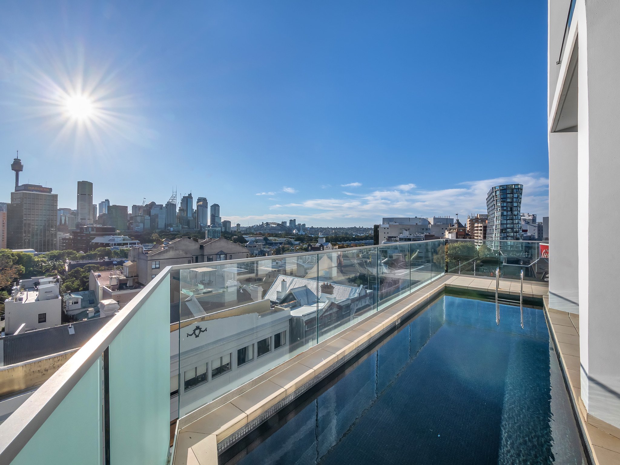 City skyline with tall buildings, a bright sun, blue sky, and an urban balcony with a swimming pool in the foreground.