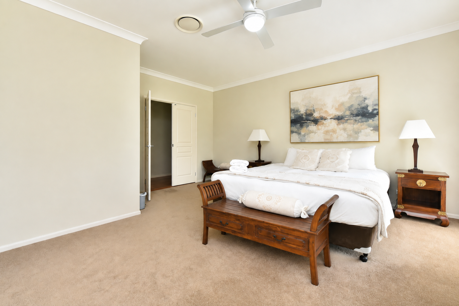 A neatly made bed with white linens in a bright hotel room, flanked by two wooden nightstands with lamps, a wooden bench at the foot of the bed, a large abstract painting above the bed, and a partially open door to the closet.