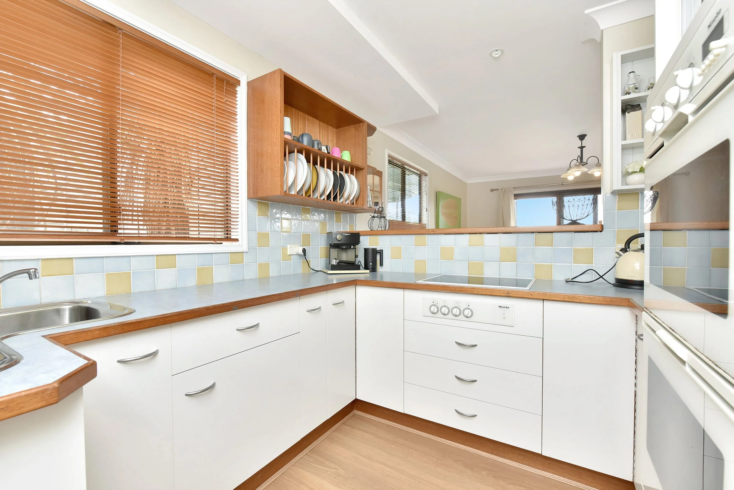 Kitchen with white cabinets, a light blue and yellow tiled backsplash, wooden countertops, and a window with brown blinds. There are shelves with dishes and a coffee machine on the counter.