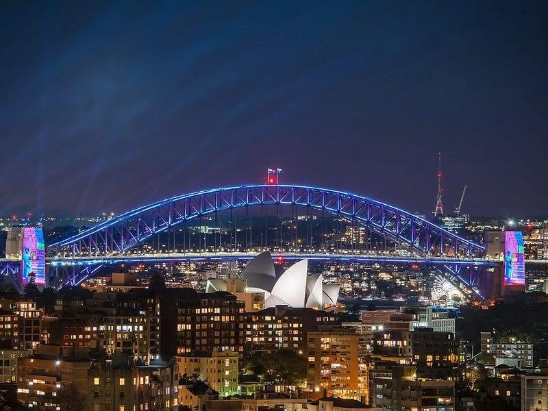 Night view of Sydney Harbour Bridge illuminated with blue lights, with the Sydney Opera House in the foreground, and city lights in the background.
