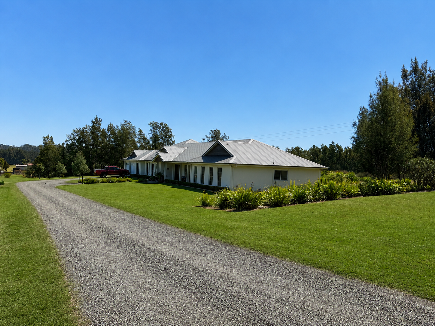 Single-story white house with a metal roof, surrounded by a well-maintained lawn and garden, situated along a curved gravel driveway under a clear blue sky.