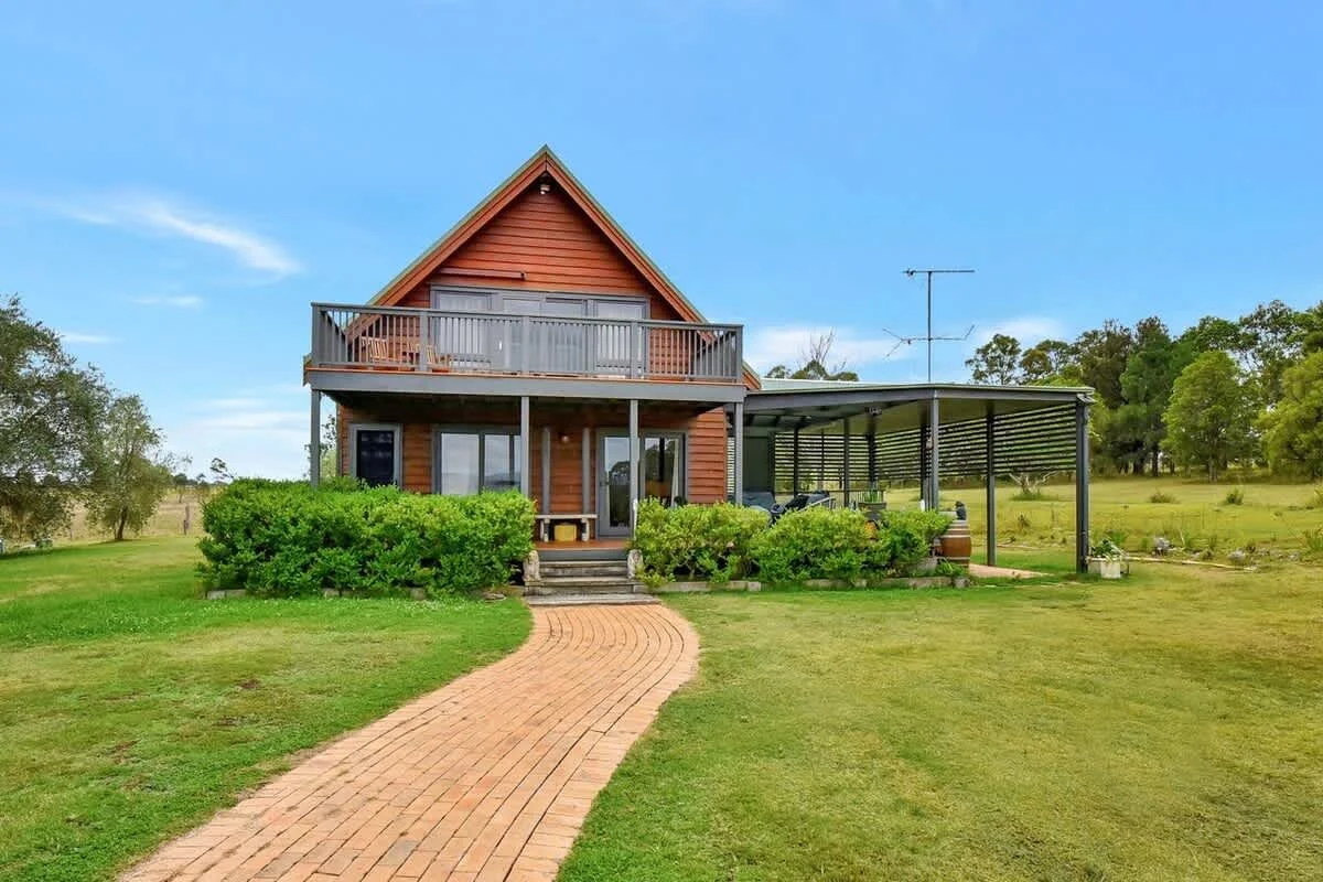 A two-story wooden house with an upper balcony and a covered patio on the ground level, surrounded by a neatly maintained lawn and bushes, with a curved brick pathway leading to the front steps, and a rural landscape with trees in the background unde
