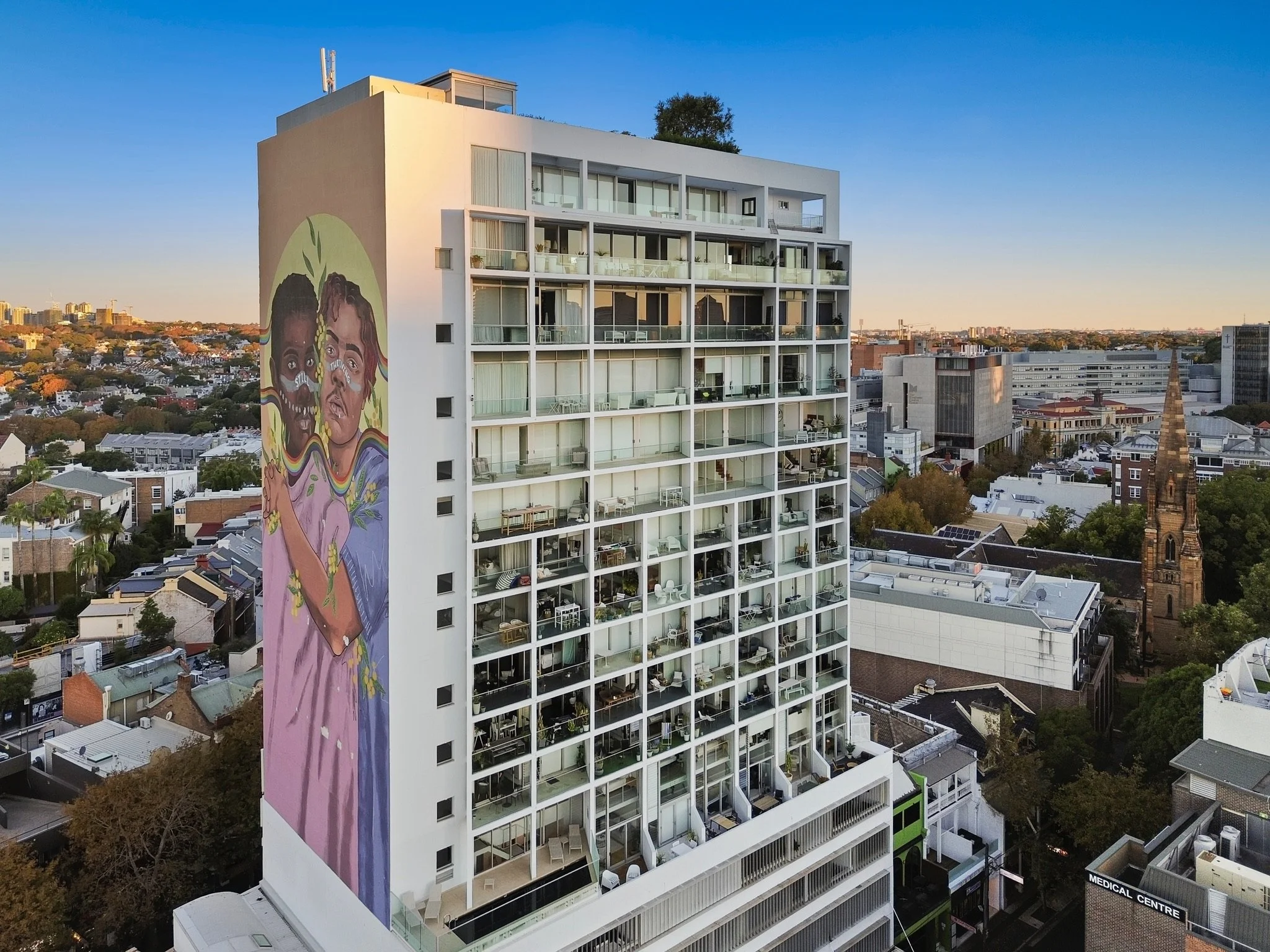 A tall white modern apartment building with a colorful mural on one side depicting two faces, one of a child and the other of a woman, with vibrant rainbow colors underneath.