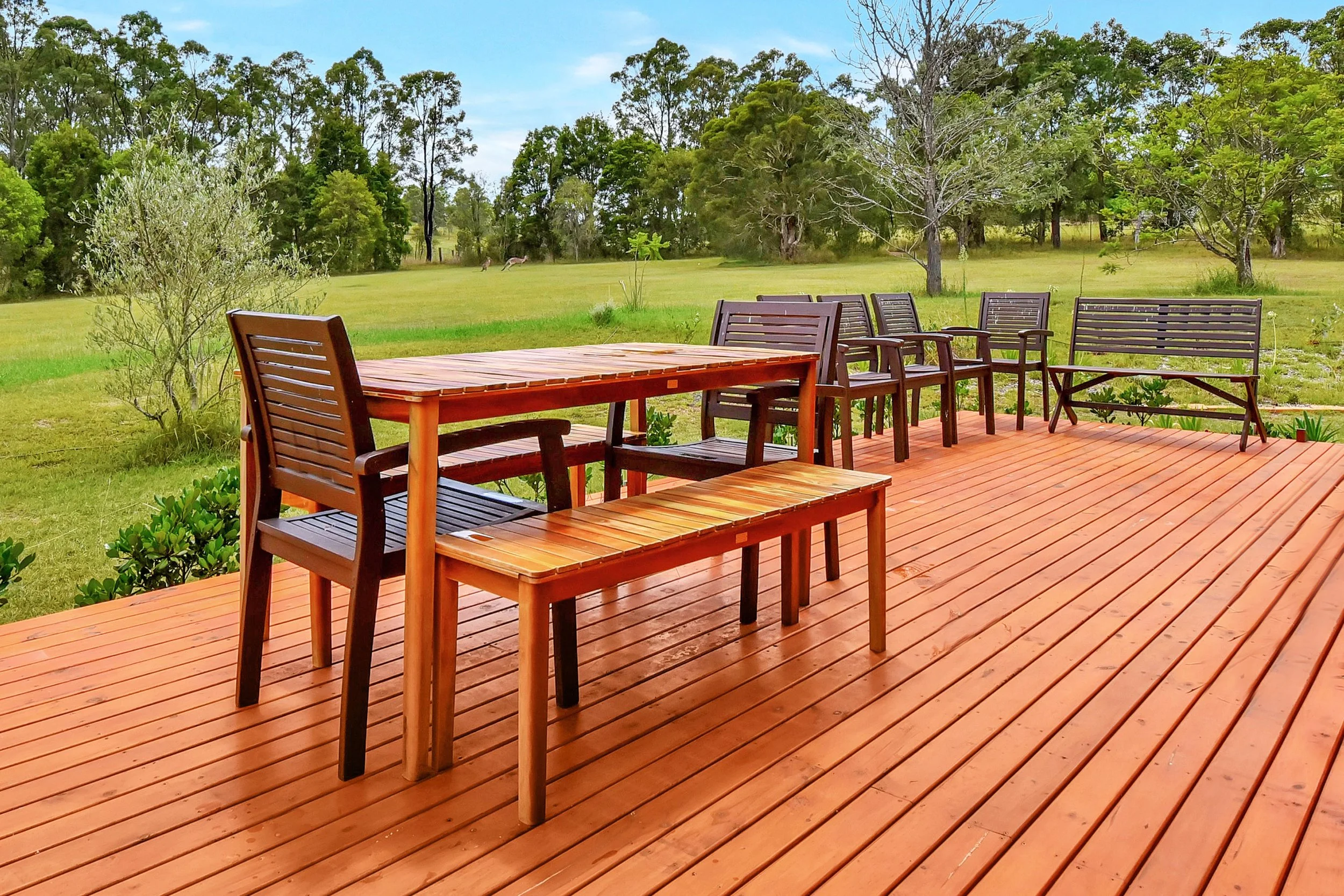 A wooden deck with outdoor furniture, including chairs and a table, overlooking a grassy field with trees and grazing animals in the background.