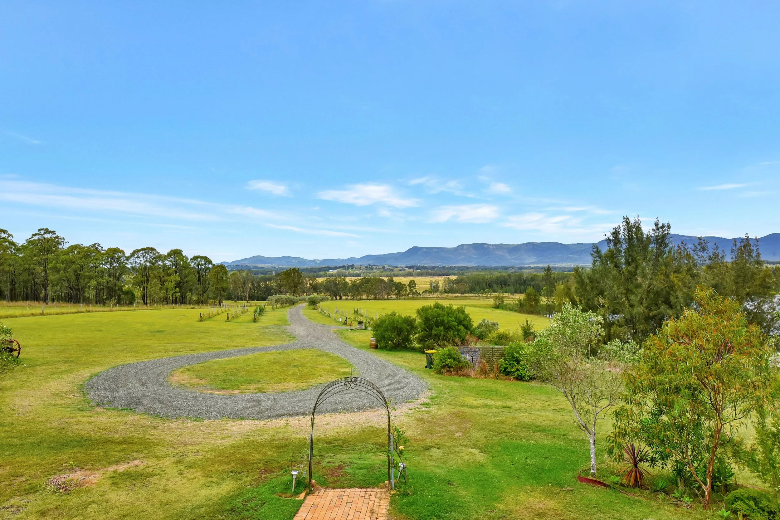 Scenic landscape with green fields, trees, a gravel path, distant mountains, and a clear blue sky.