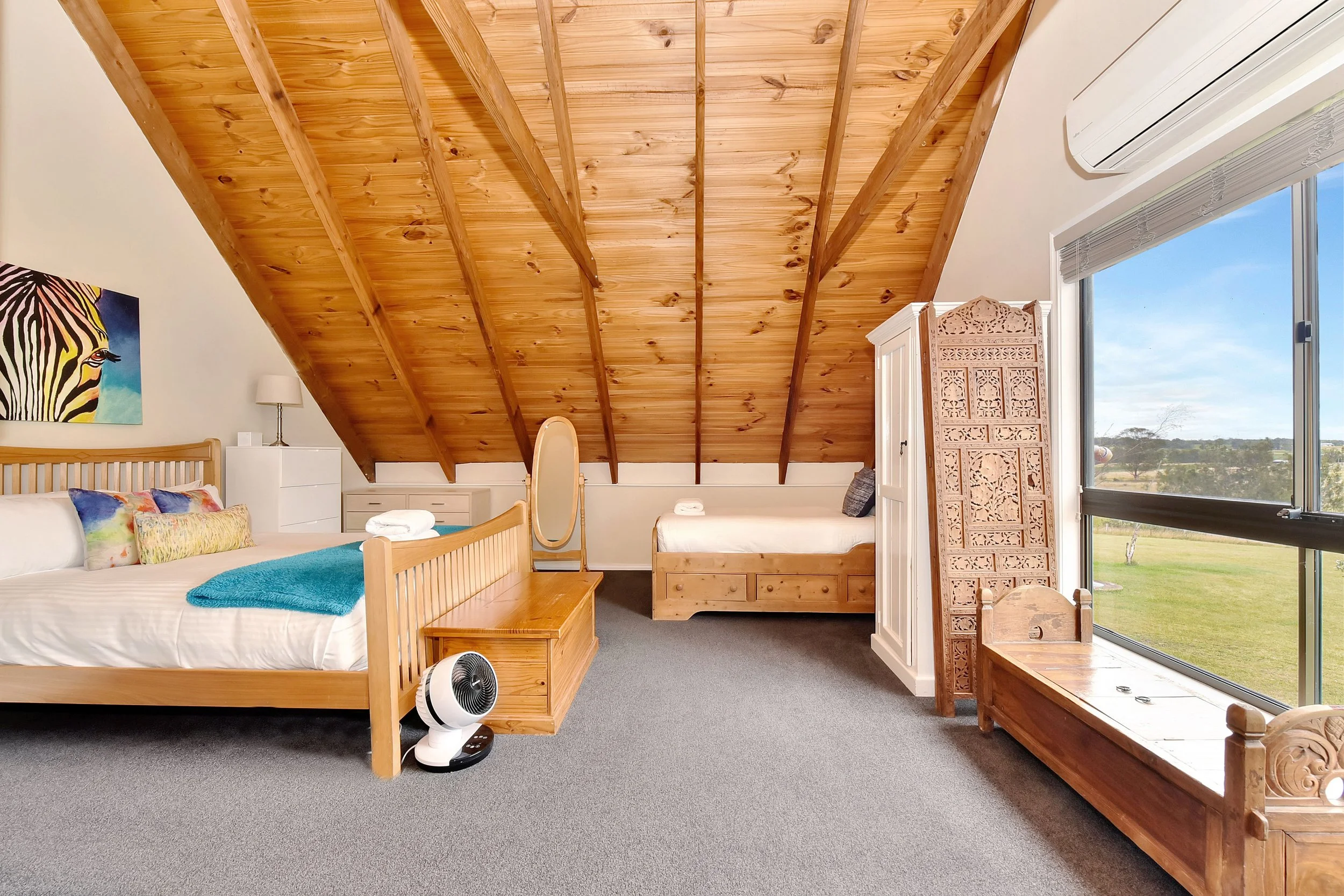 A cozy bedroom with a sloped wooden ceiling, two beds, a wooden bench at the foot of the bed, and a large window showing a rural landscape.