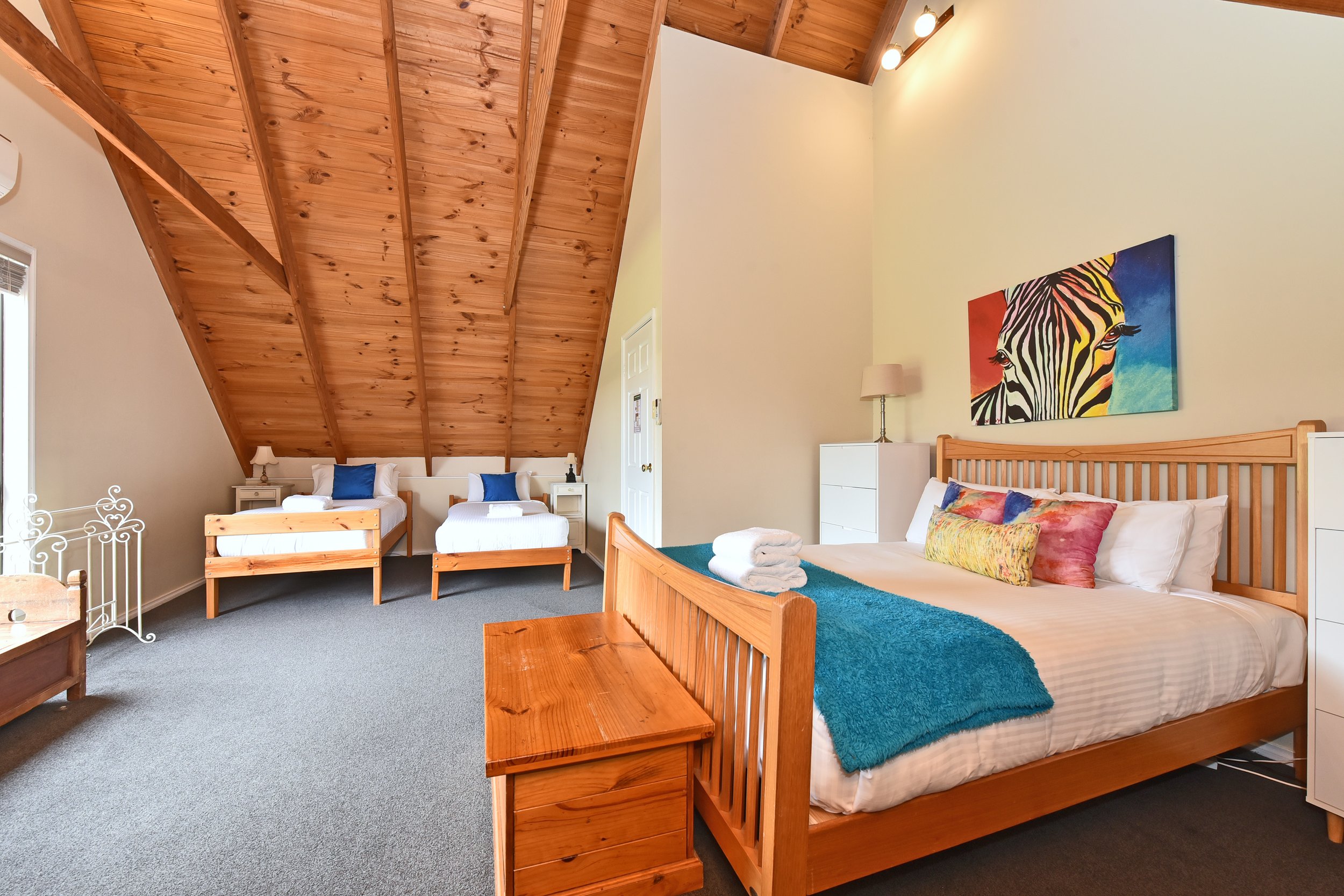 A bedroom with two beds with wooden frames, colorful pillows, and a turquoise blanket on the larger bed. There is a wooden nightstand with a lamp and a piece of colorful art of a zebra on the wall. The ceiling has exposed wooden beams.