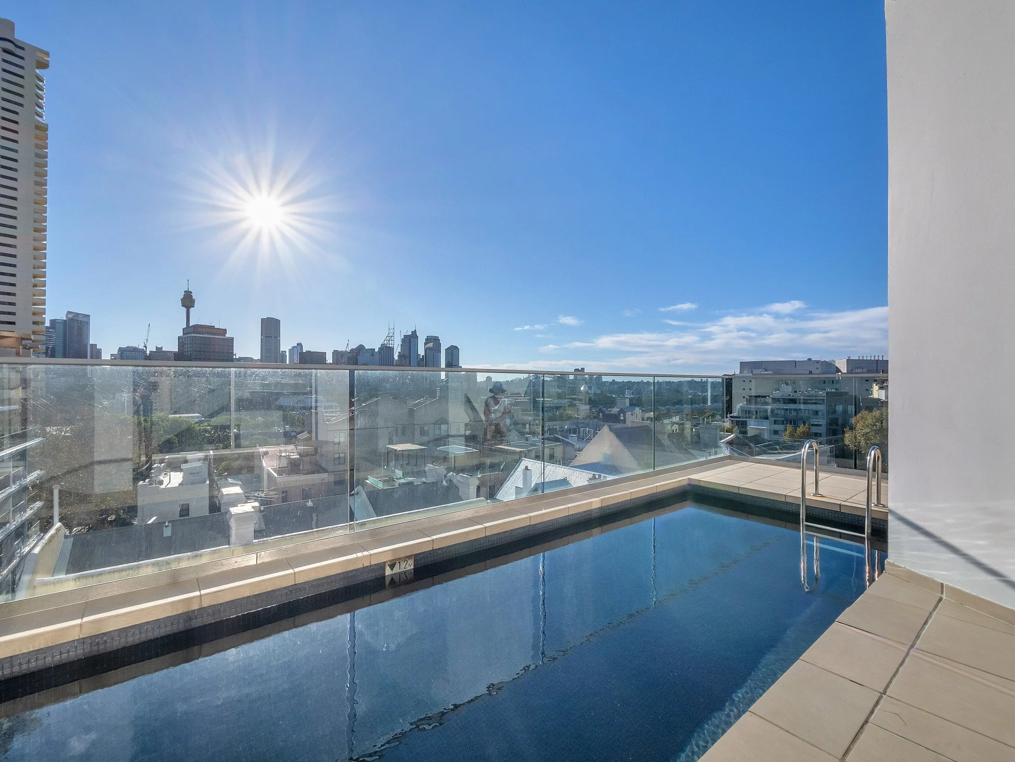 Urban city skyline with tall buildings and a tower with an observation deck, seen from a rooftop pool area under a clear blue sky with the sun shining brightly.