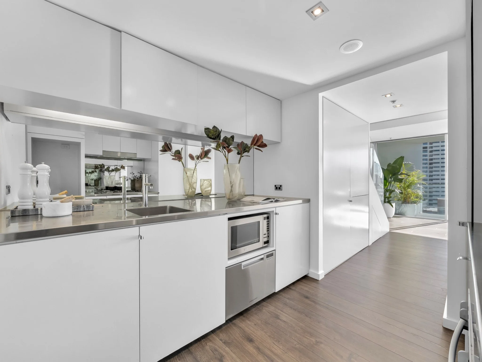 Modern white kitchen with a stainless steel sink, a microwave, decorative vases, and a large window with a city view.