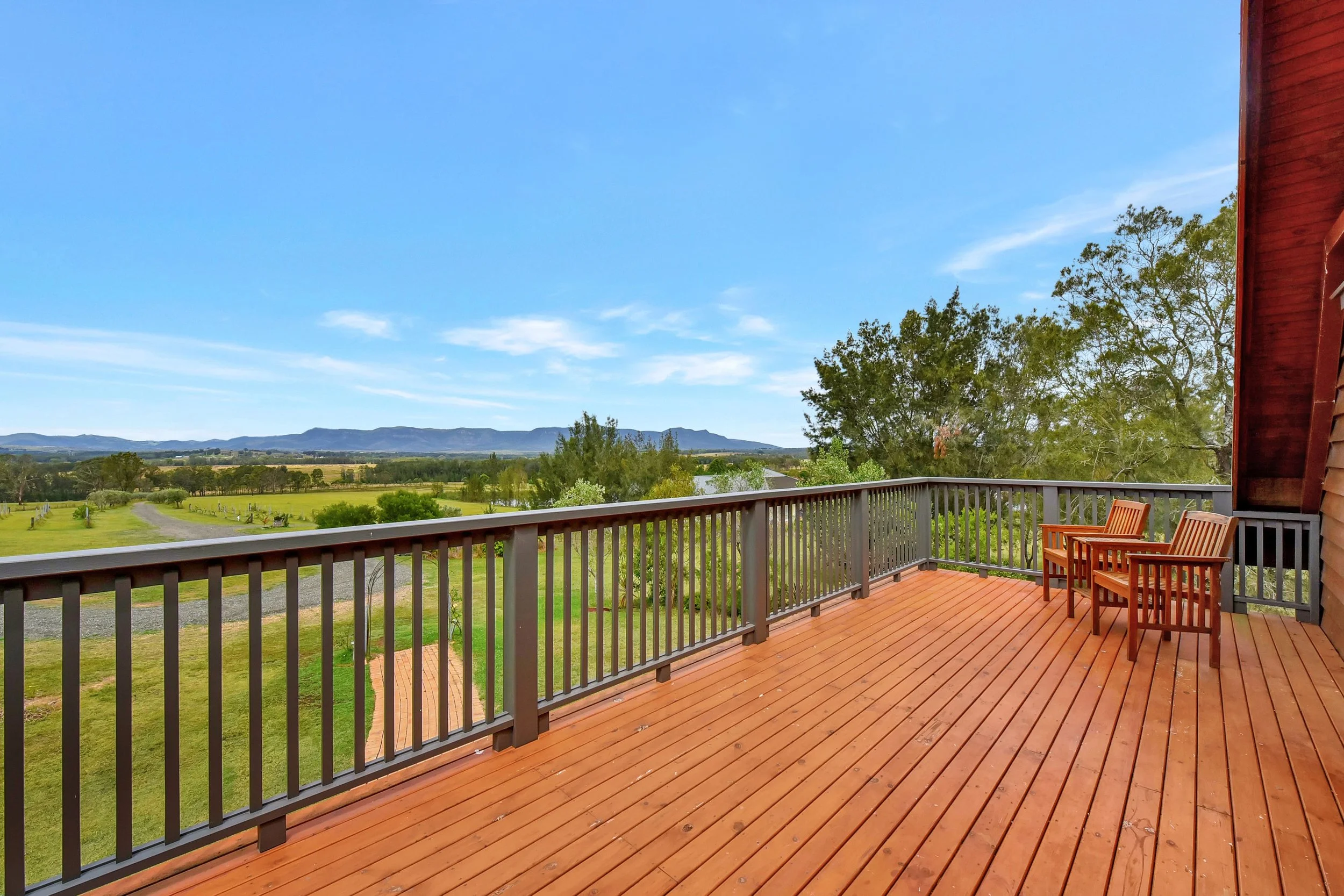 A wooden deck with chairs overlooking green fields, trees, and distant mountains on a sunny day with blue sky