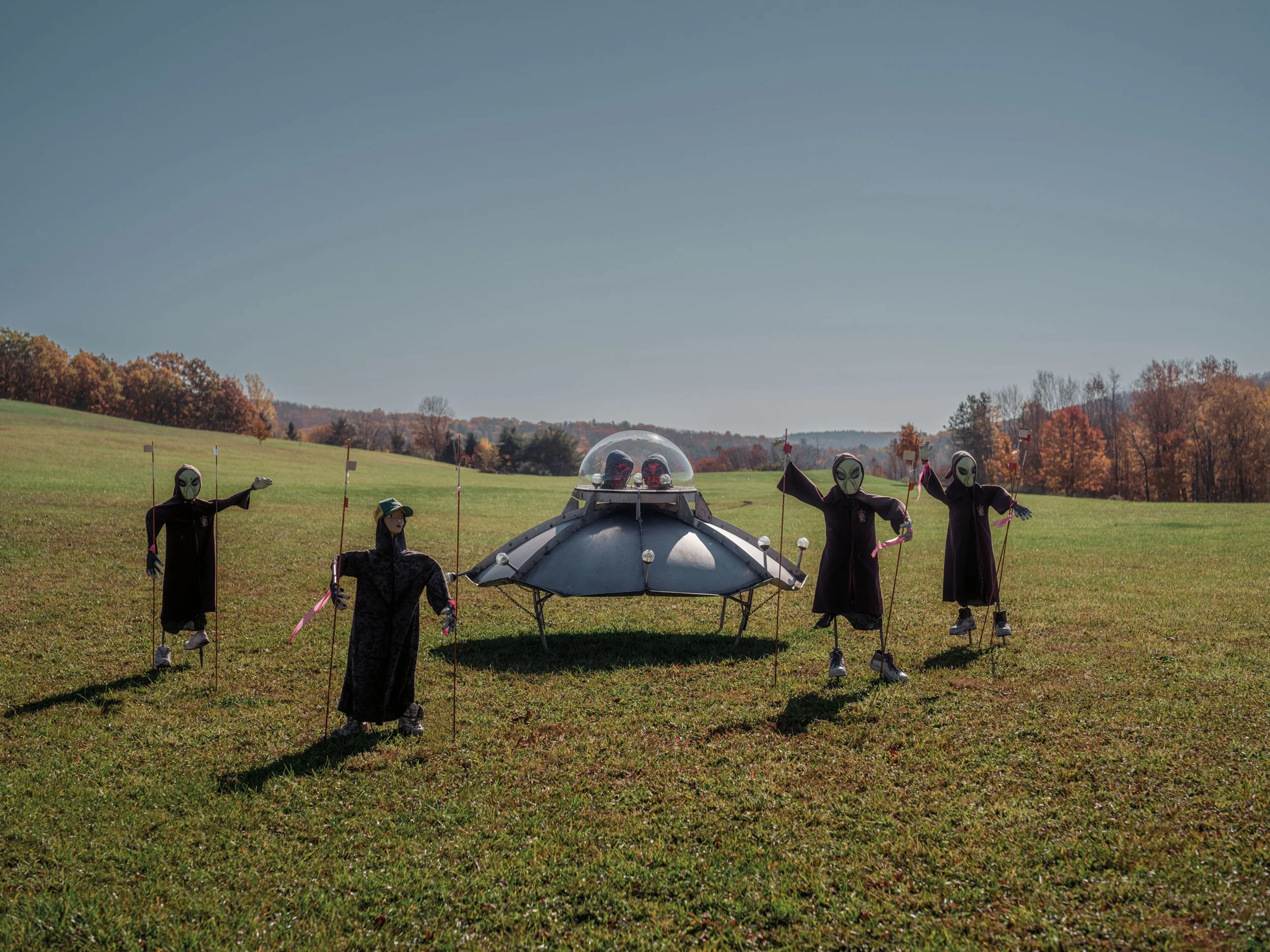 A group of people dressed in black with alien masks and clothes, holding sticks, standing near a small vehicle with a dome-shaped top, on a grassy field with autumn-colored trees in the background.
