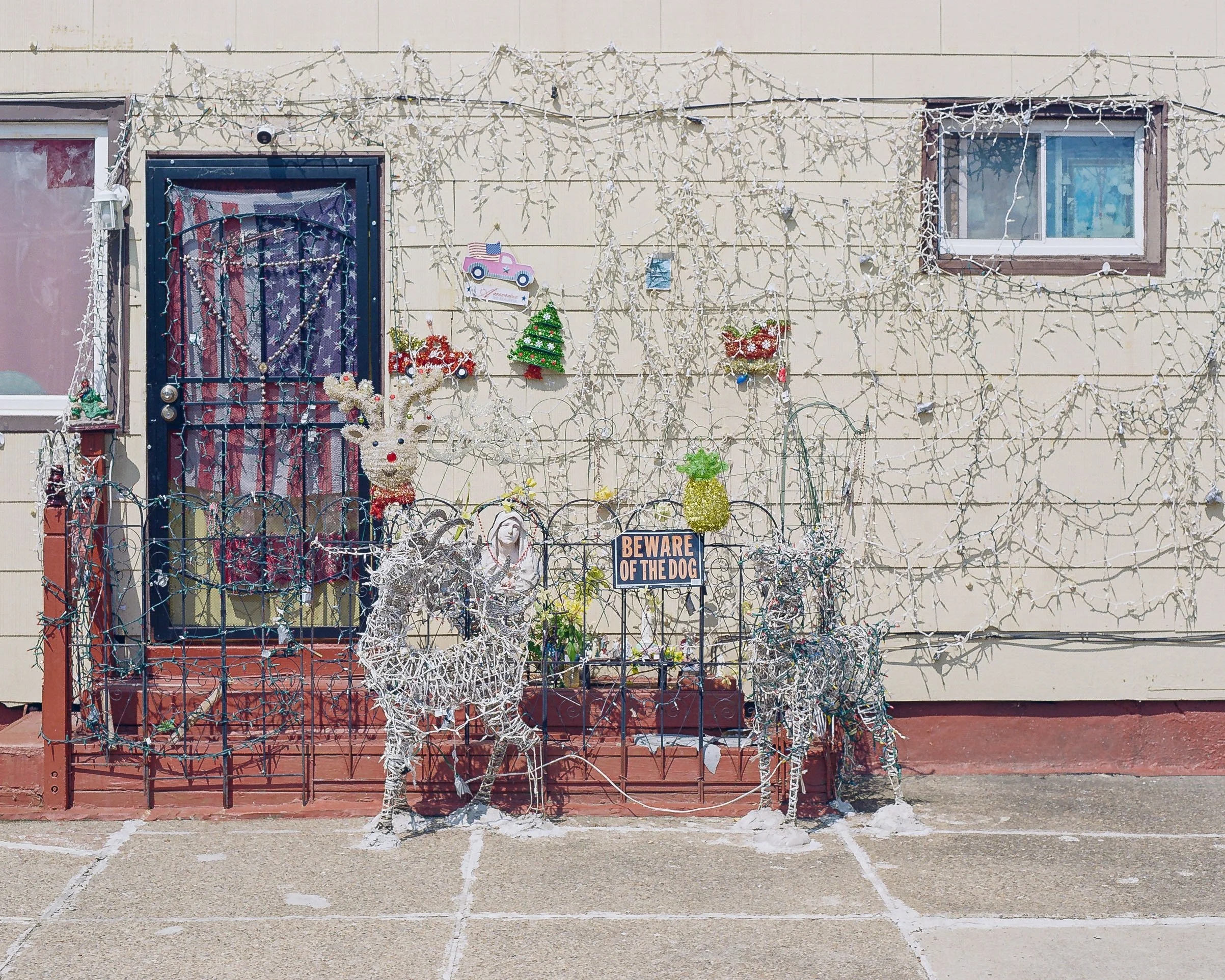 A house exterior decorated with white string lights, holiday ornaments, a reindeer decoration, and a sign that says 'Beware of the Dog.' The house has beige siding, a window, and a door with a decorative security screen.