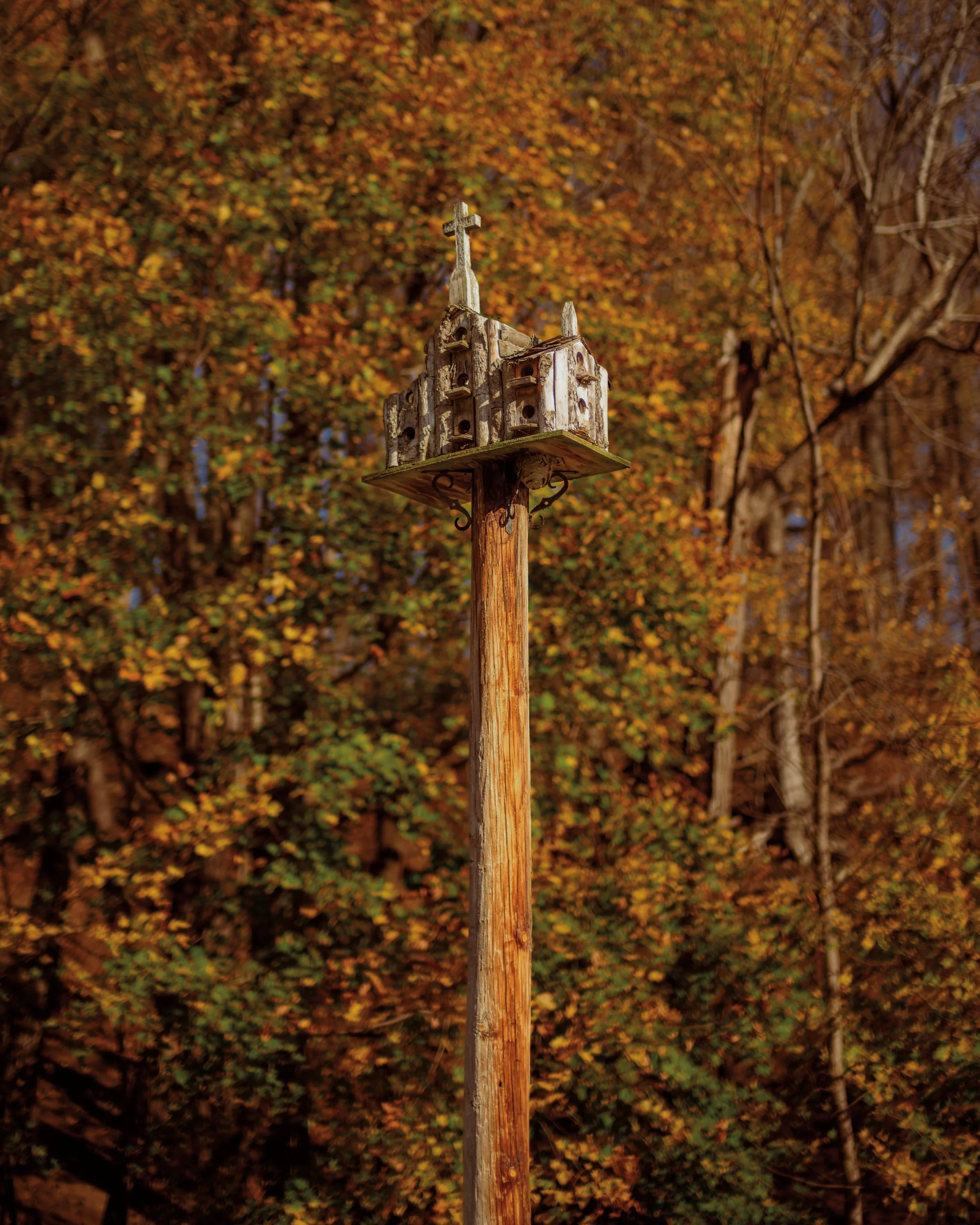 An old wooden birdhouse resembling a church with a cross on top, mounted on a pole with decorative metal brackets, set against a background of colorful fall foliage.