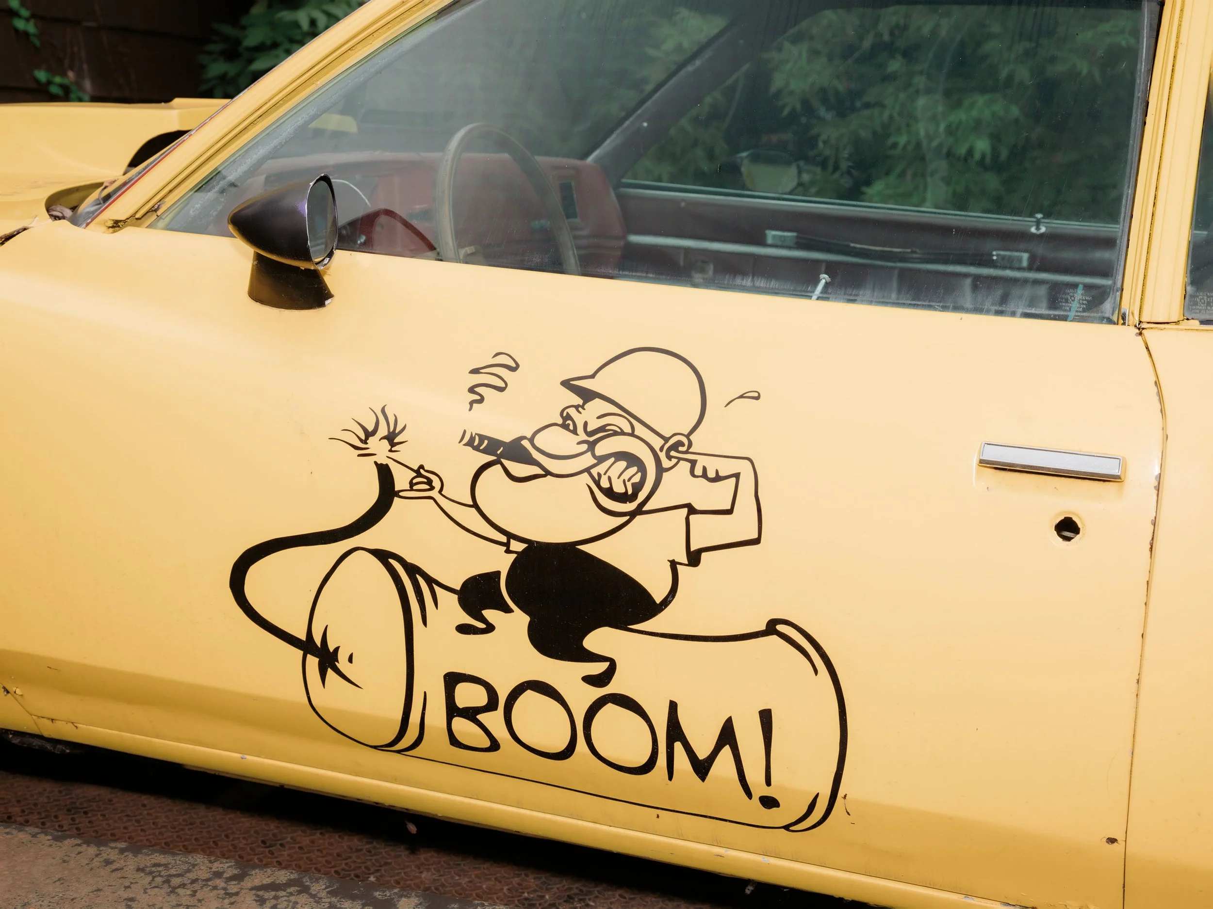 Yellow car with a cartoon sticker of a person with a hat, smoking and holding a lit fuse, and the word 'POW' written below.