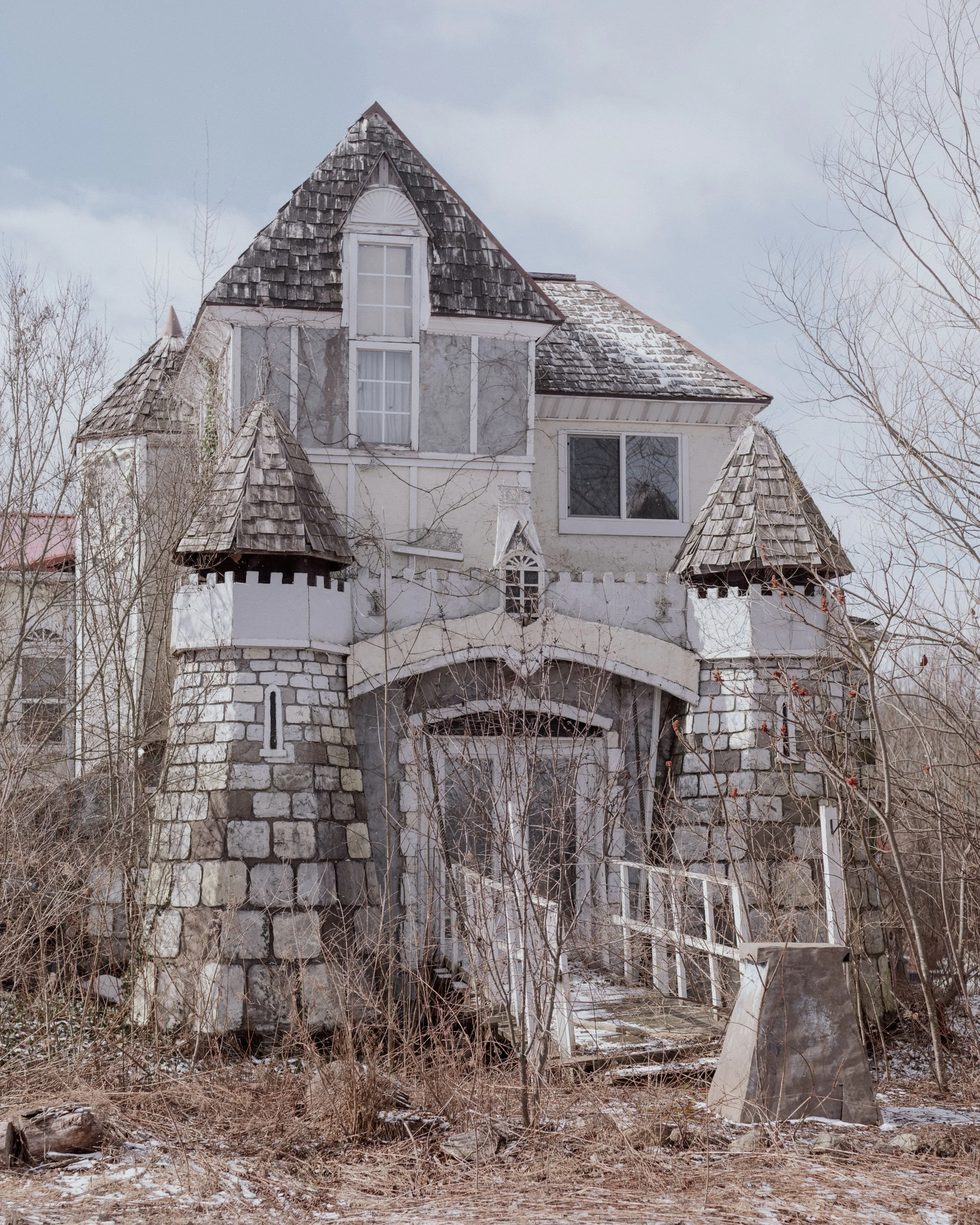 Old, weathered house with steep roof, multiple gables, and a stone foundation, surrounded by leafless trees and overgrown vegetation.