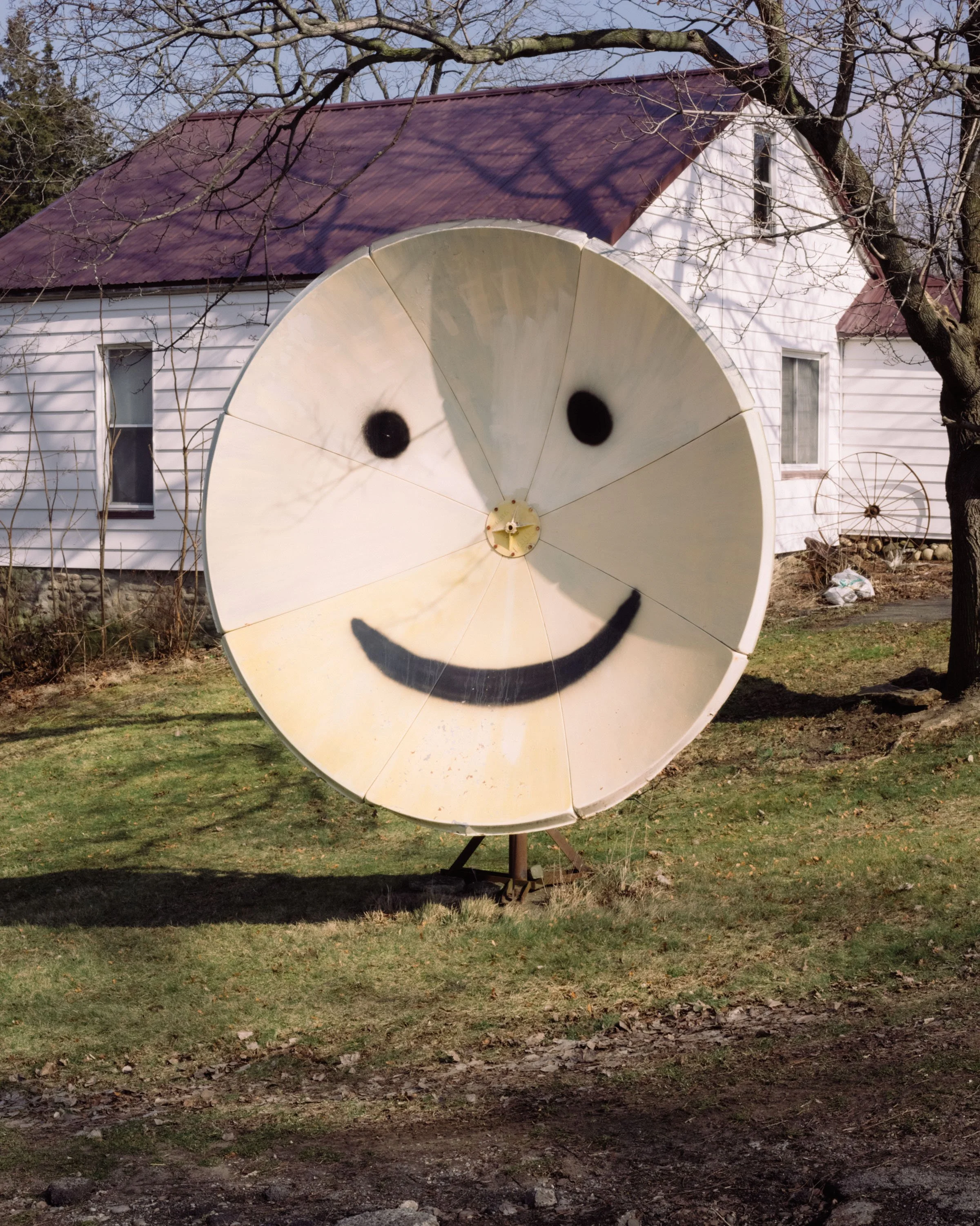 A large satellite dish with a smiley face drawn on it, situated on a grassy yard with a white house and bare trees in the background.