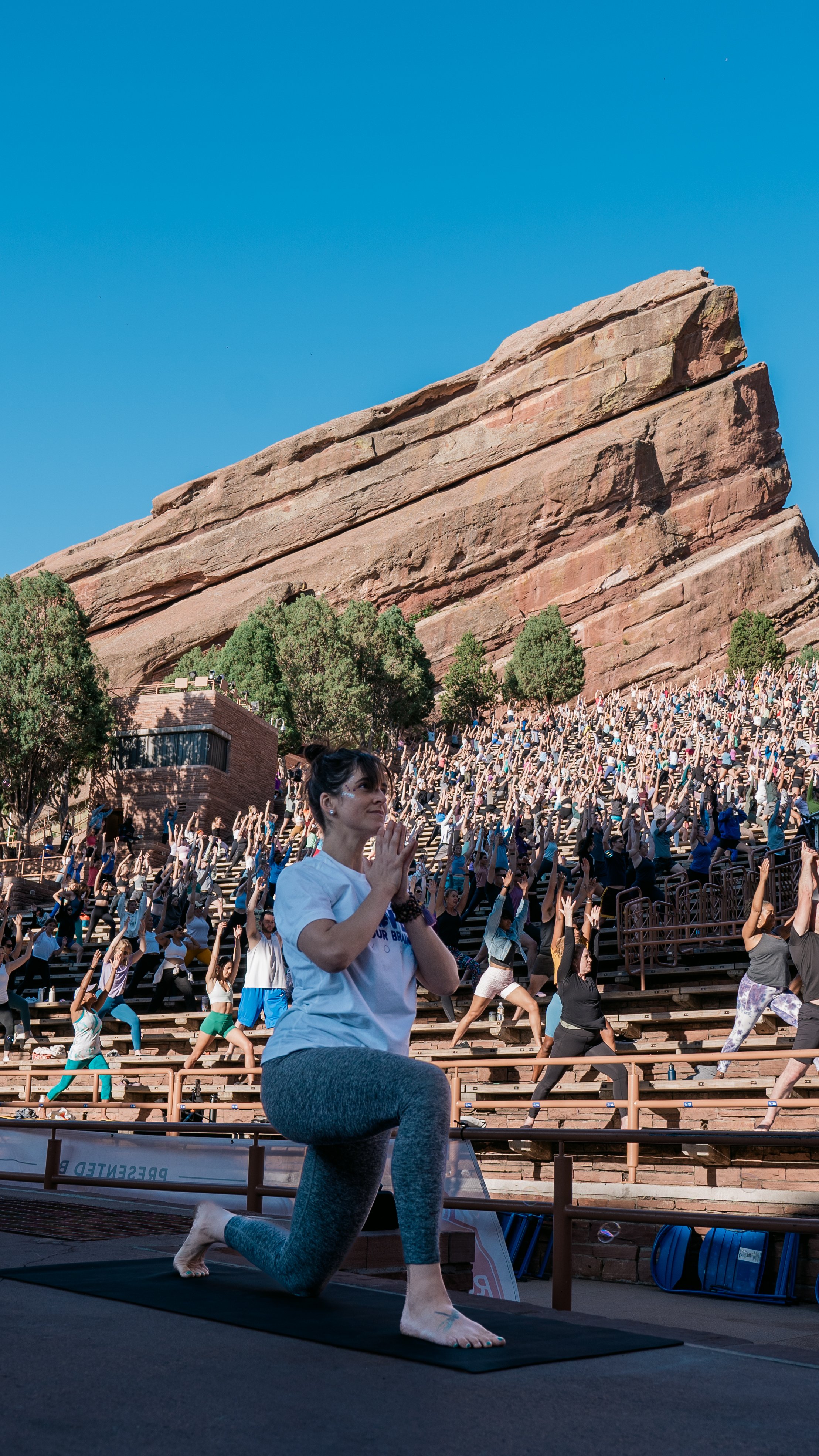 A woman practicing yoga and meditation on a black yoga mat on bleachers during an outdoor event, with a large group of people doing yoga in the background against a rocky hillside and a clear blue sky.