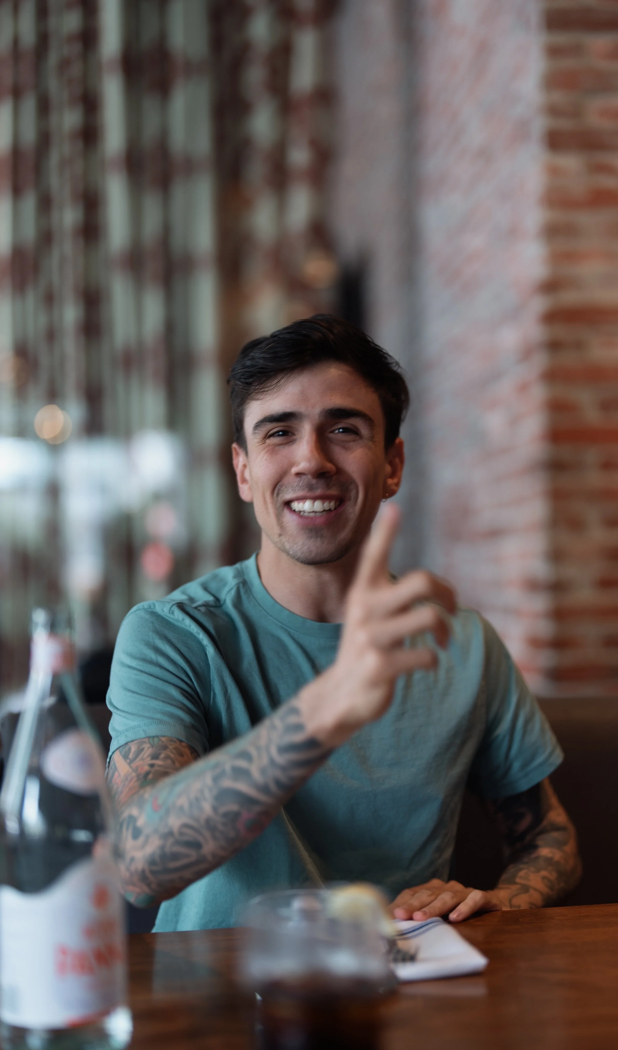 Young man with dark hair and tattoos on his arms smiling and pointing his finger, sitting at a dining table in a restaurant with a brick wall and glass behind him.