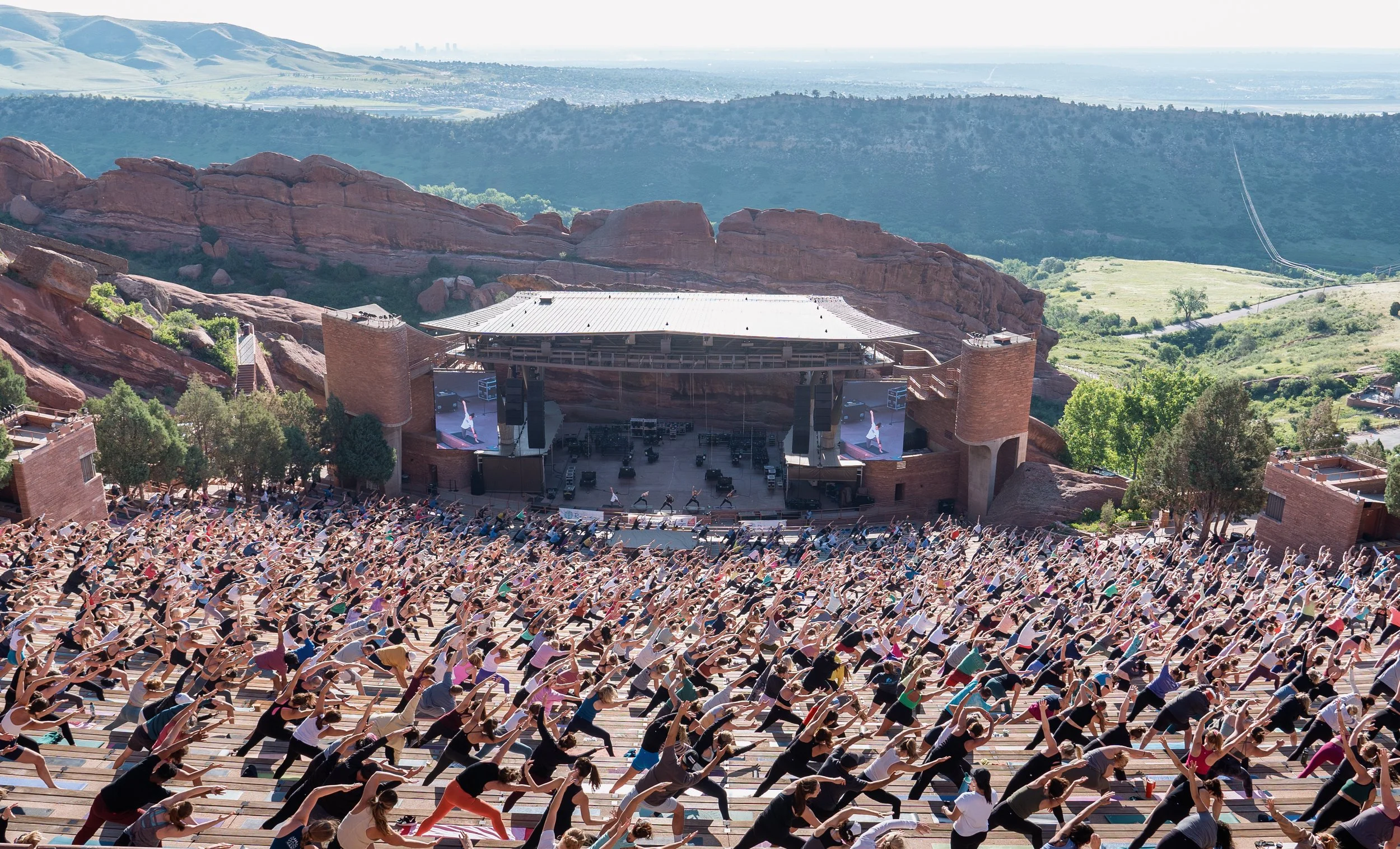 A large outdoor yoga class with hundreds of participants practicing on wooden steps, set against a mountainous landscape with red rocks and scenic distant hills.