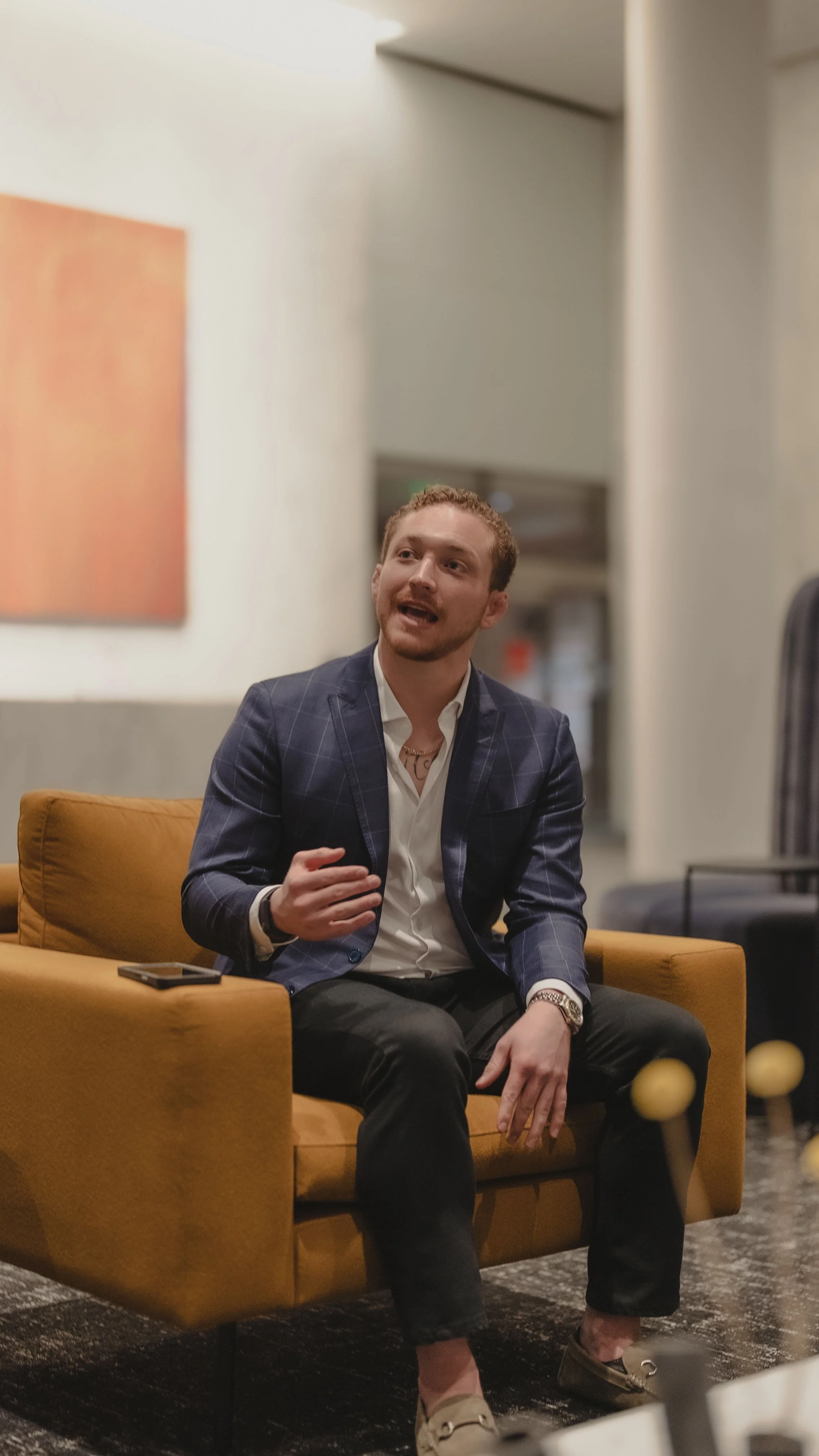 A young man with reddish hair and a beard, dressed in a dark checkered blazer, white shirt, and black pants, is seated on a mustard-colored sofa, speaking and gesturing with his hands. There is a smartphone on the armrest next to him, and the background shows a softly lit room with neutral-colored walls.