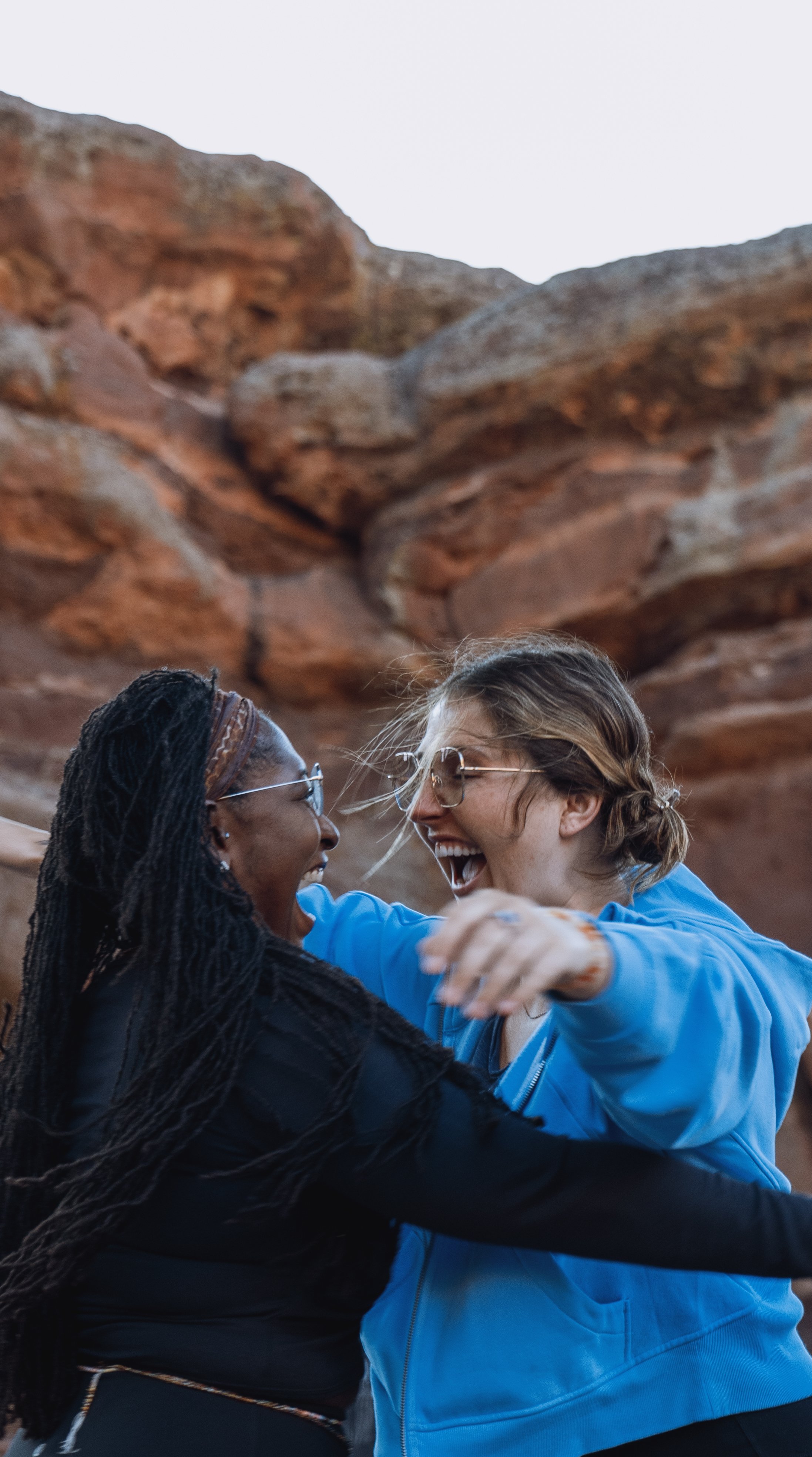 Two women with glasses and wide smiles, embracing each other outdoors against a rocky background.