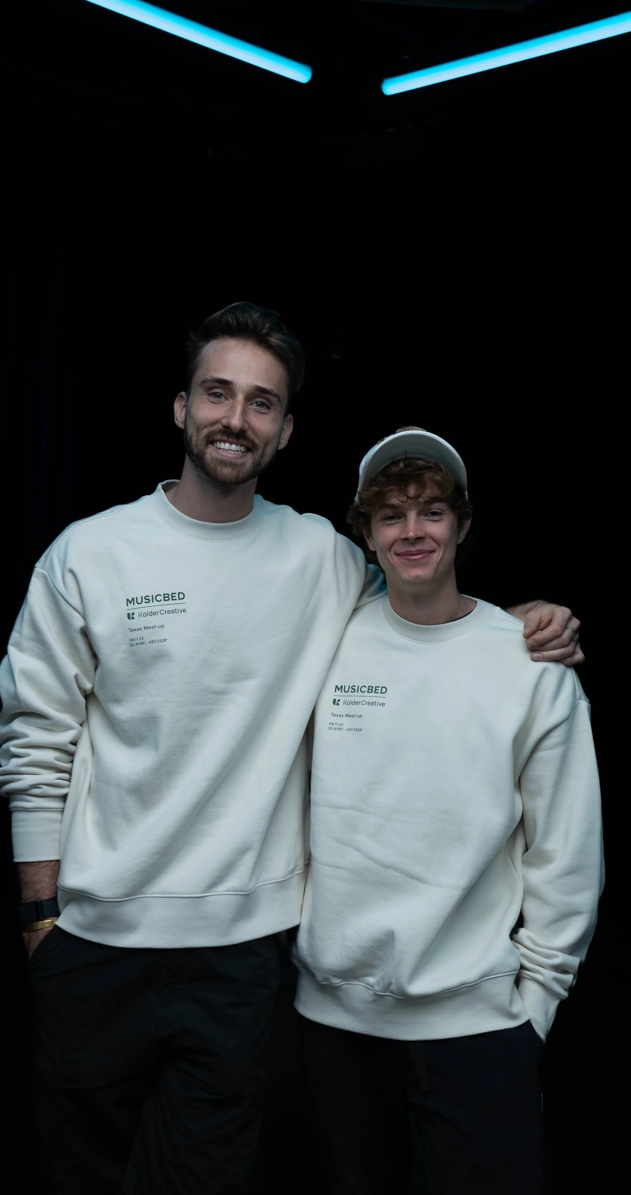 Two young men smiling with arms around each other, wearing matching cream-colored sweatshirts with 'MUSICBED' printed on them, standing against a black background with blue neon lights overhead.