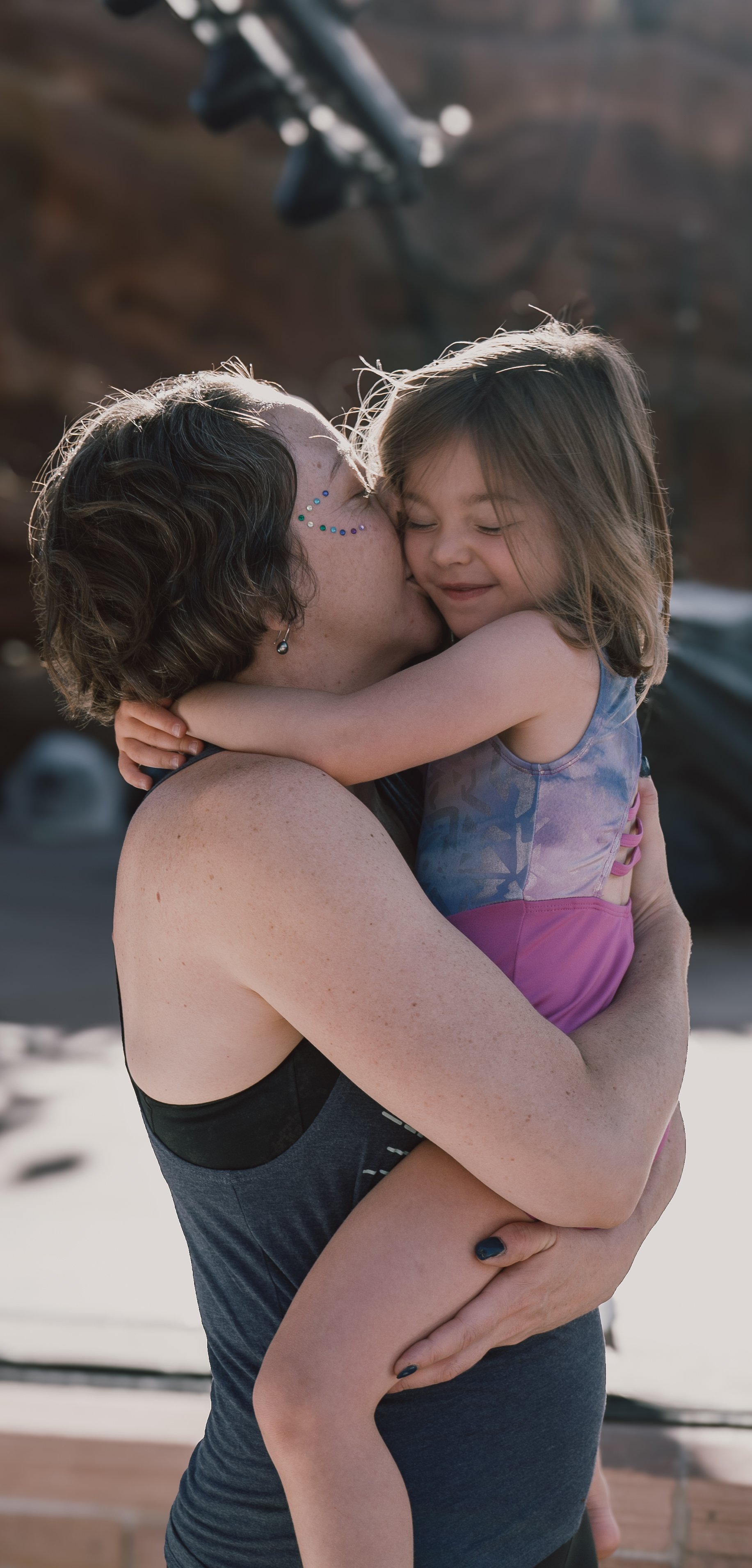A woman with short hair and face rhinestones is holding a young girl with long hair, wearing a colorful swimsuit, in a hug. They are outdoors in bright sunlight.