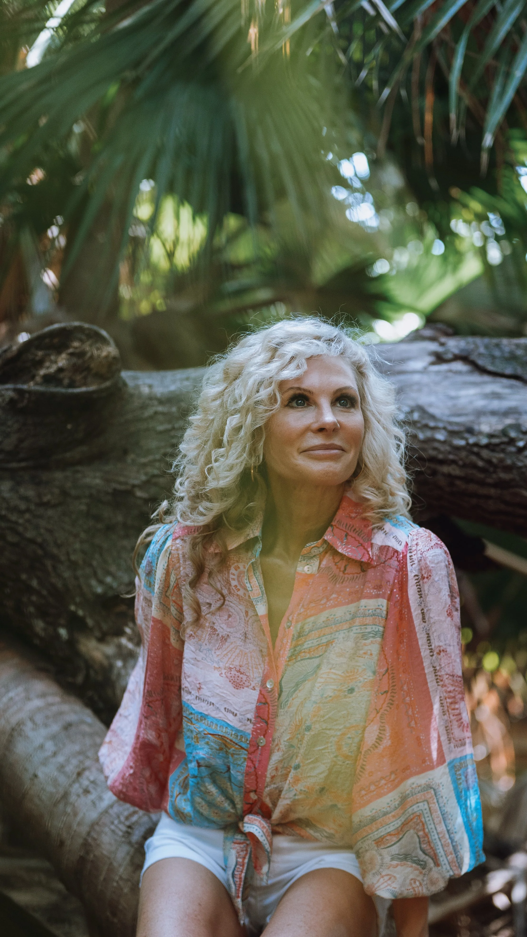A woman with curly blonde hair, wearing a colorful patterned shirt and white shorts, sitting on a fallen tree trunk in a lush green forest.