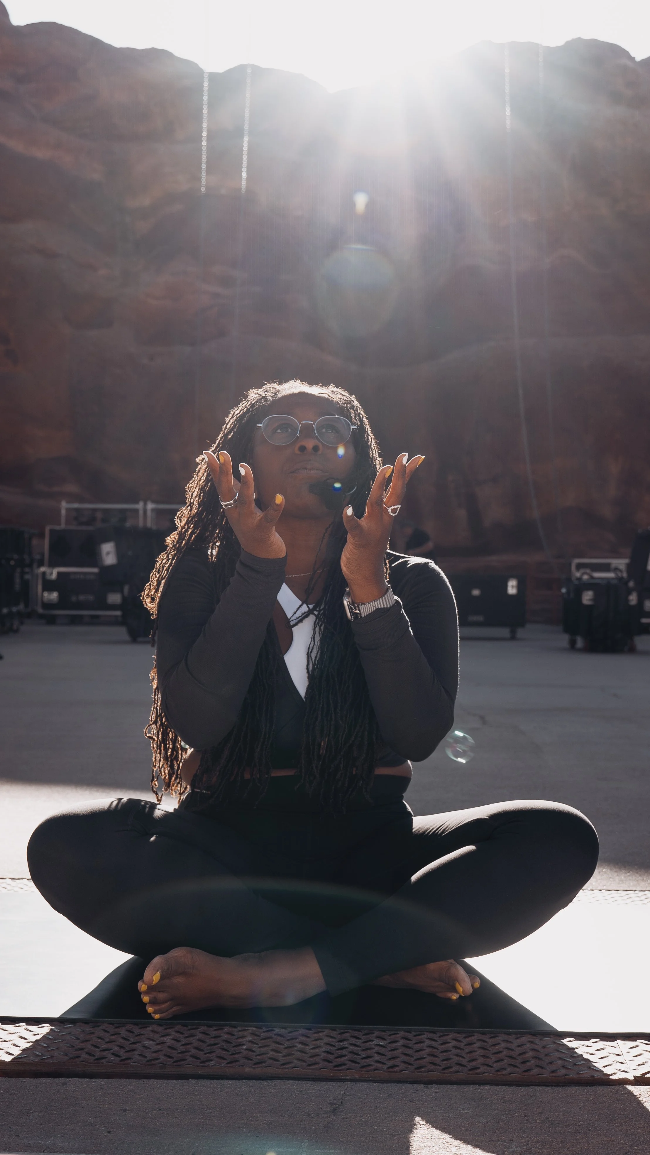 A woman with glasses sitting cross-legged on the ground with her hands raised near her face, in a rocky outdoor setting with a canyon wall behind her and bright sunlight shining overhead.