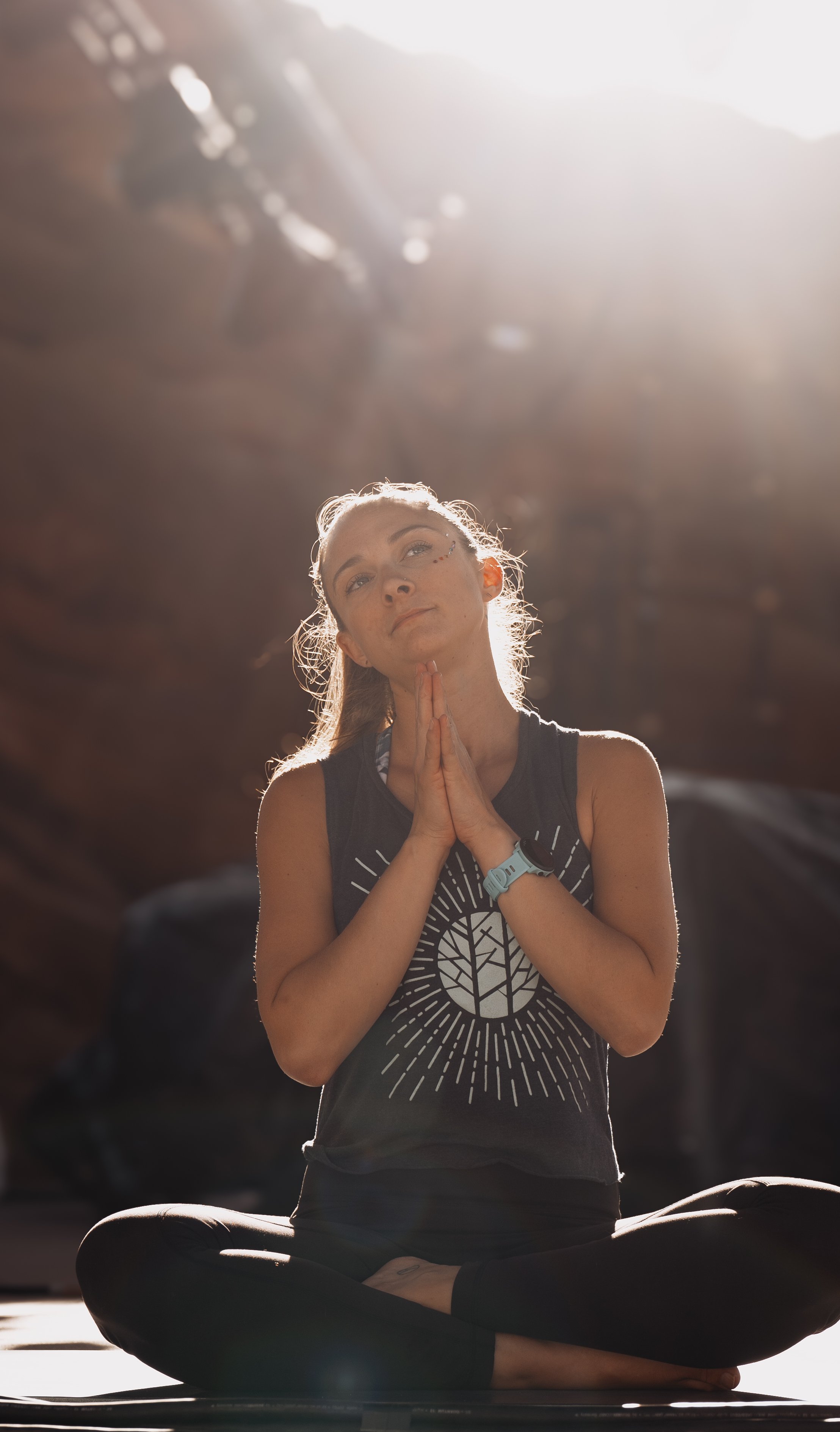 A woman sitting cross-legged on the floor, meditating with her hands in prayer position in front of her chest, under soft sunlight with a blurred background.