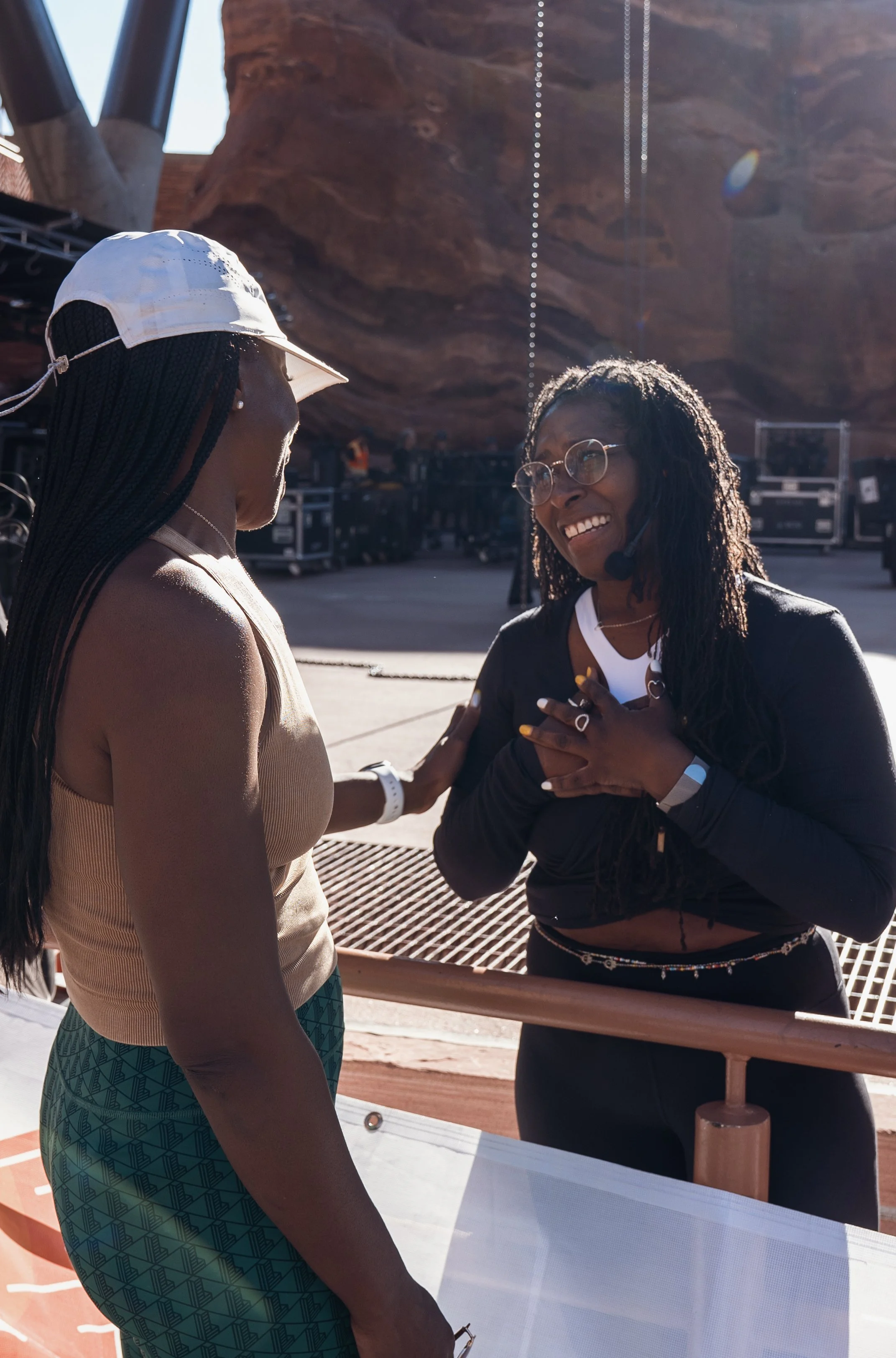 Two women interacting outdoors at a rocky location, one wearing a white cap and tank top, and the other with glasses and a microphone headset, both smiling and engaged in conversation.