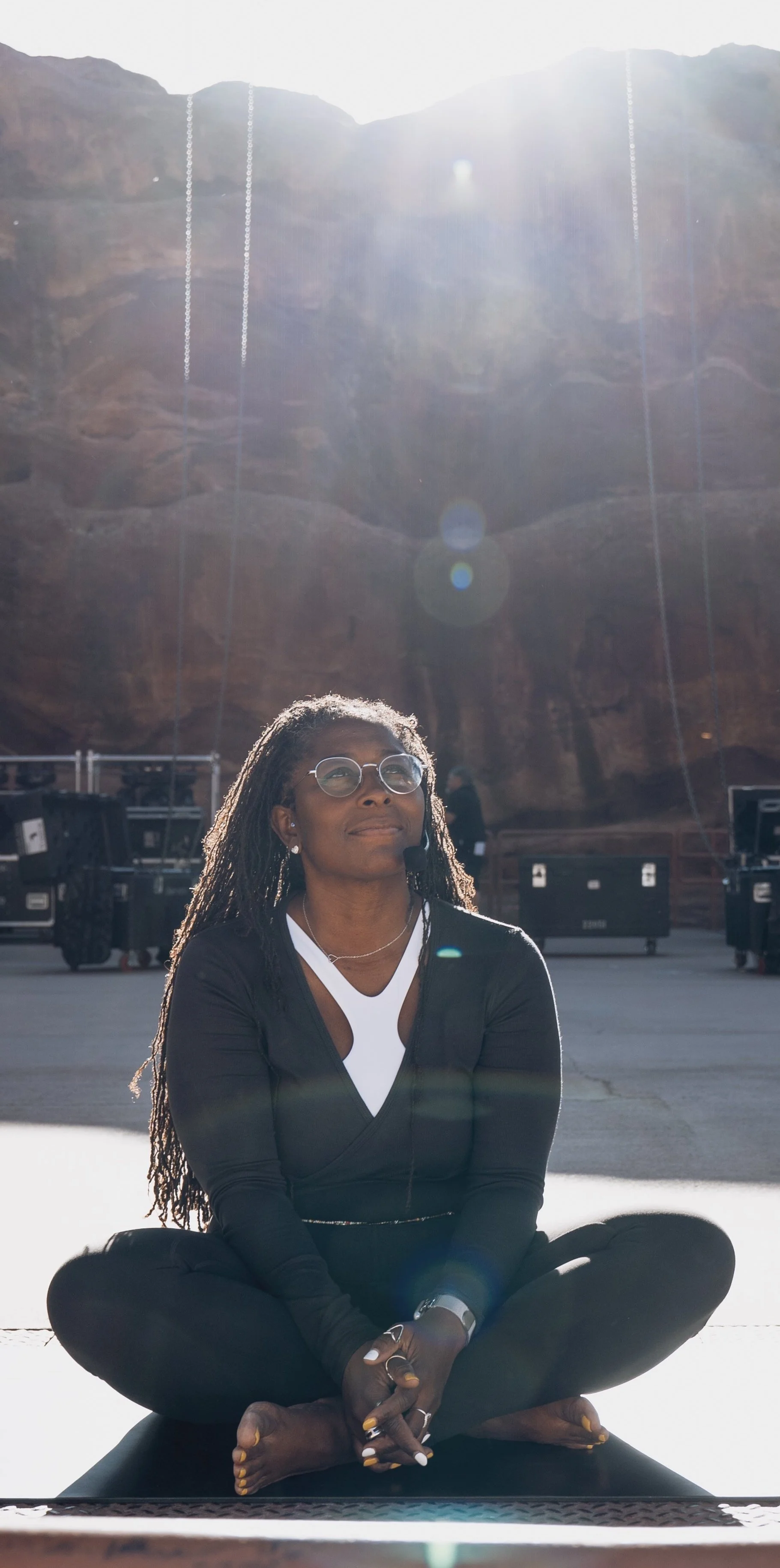 A woman practicing yoga outdoors at the base of a canyon with bright sunlight overhead, sitting cross-legged on a yoga mat, wearing glasses and a microphone headset.