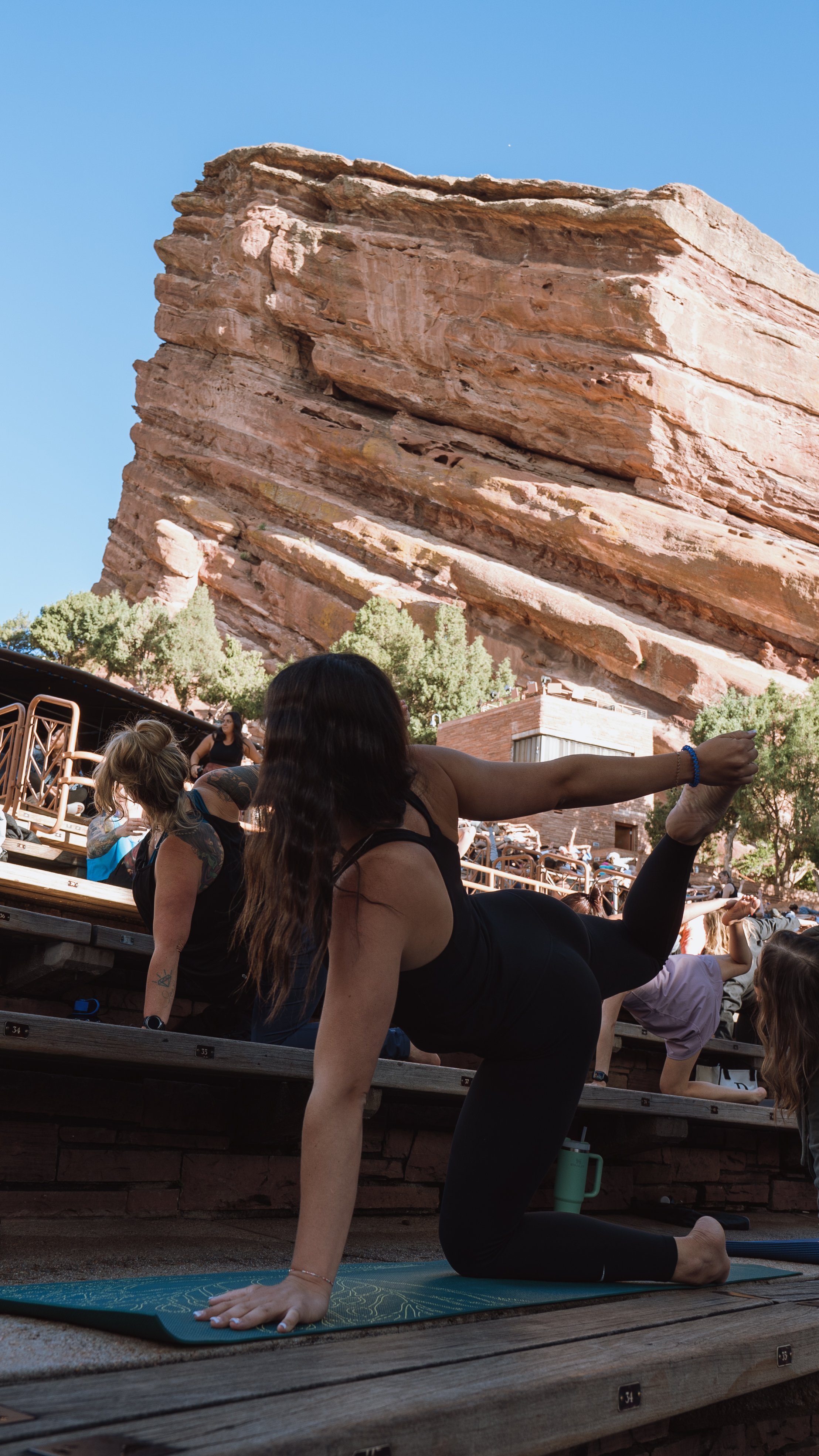 A woman practicing yoga outdoors on a wooden deck with a large rock formation and trees in the background, performing a yoga pose on a mat.