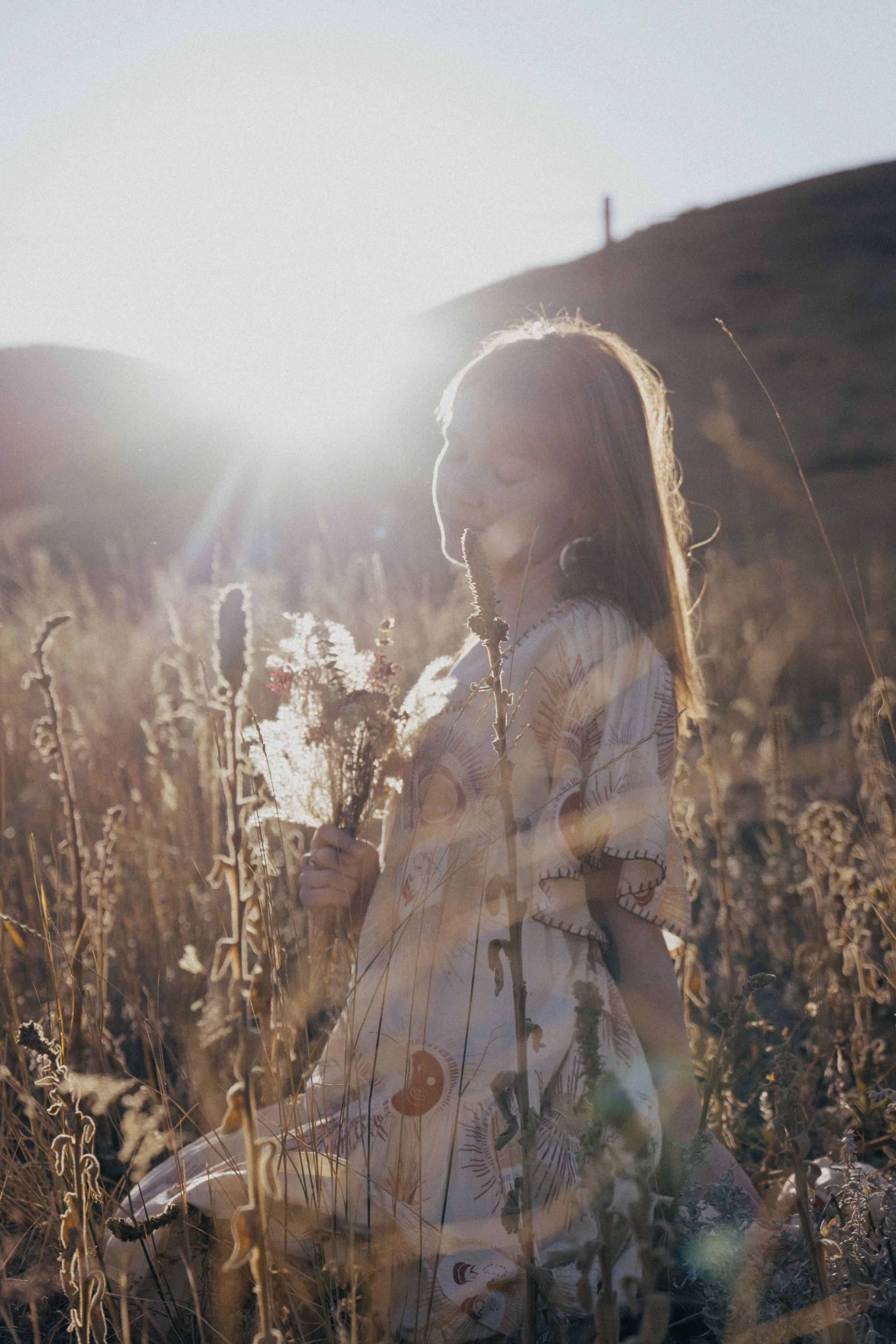 A woman with long hair standing in a field of tall, dry plants during sunset, wearing a patterned dress and holding a bouquet of flowers.