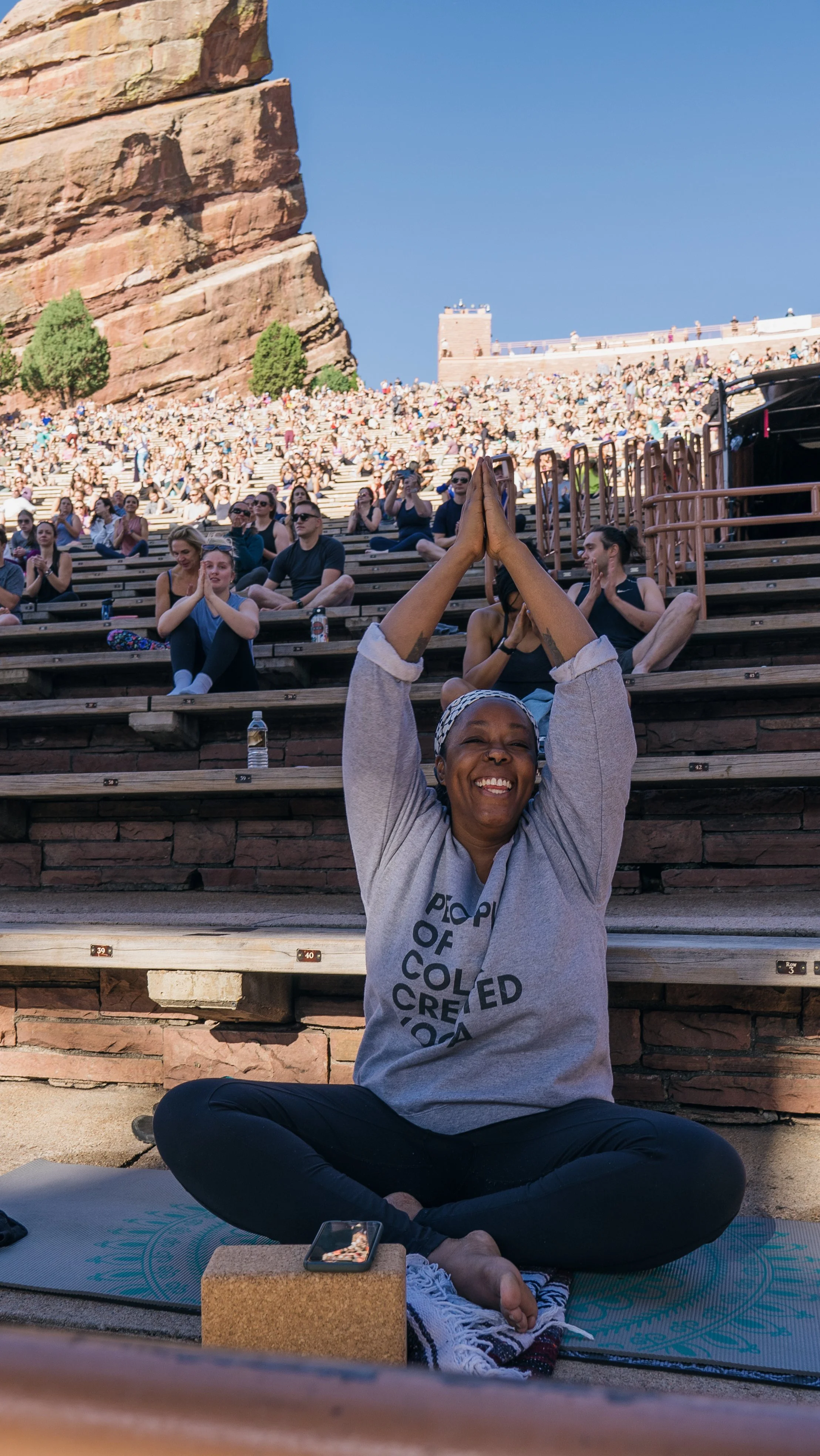 Person sitting cross-legged and smiling, practicing yoga or meditation outdoors with a cityscape and staircase filled with large crowd behind her, during daytime.