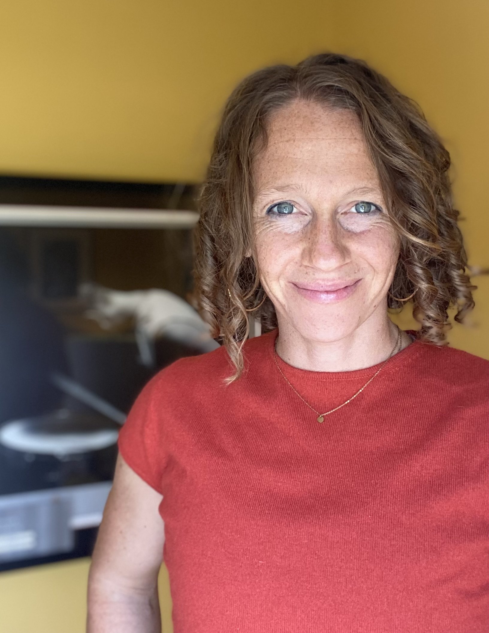 Burnout recovery coach with curly hair smiling, wearing red shirt against yellow background.